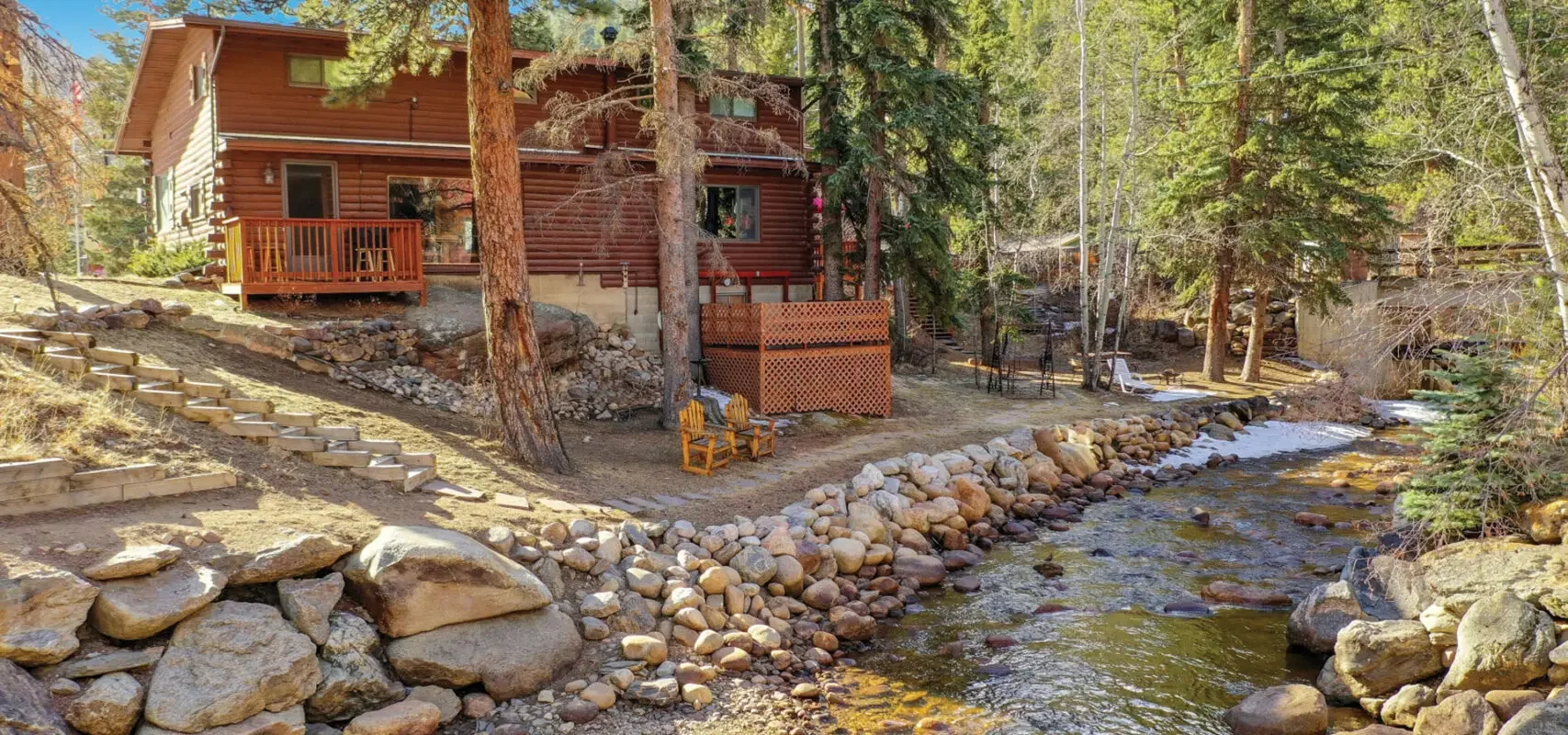 A wooden house with a backyard beside a flowing creek, surrounded by trees, with outdoor chairs and a stone pathway.