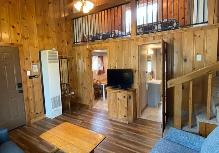 Interior of a cozy wood-paneled living room with a small wooden coffee table, a flat-screen TV on a wooden stand, a staircase, and a loft area with beds visible.