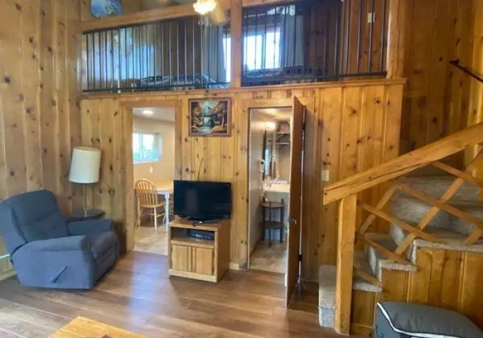 Interior view of a cozy wooden cabin living room with a blue armchair, a standing lamp, a small TV on a wooden stand, and a staircase leading to an upper level. There's a door to a bathroom and a doorway to a dining area with a wooden table and chairs. A painting decorates the wooden paneled wall.