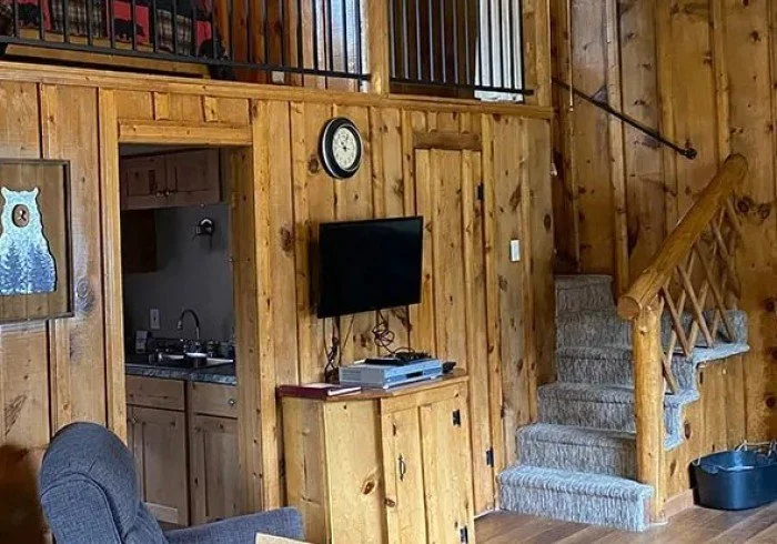 Wood-paneled living room with a staircase, a small cabinet with a TV and a DVD player, a wall clock, and a door leading to a kitchen with a sink and cabinets.