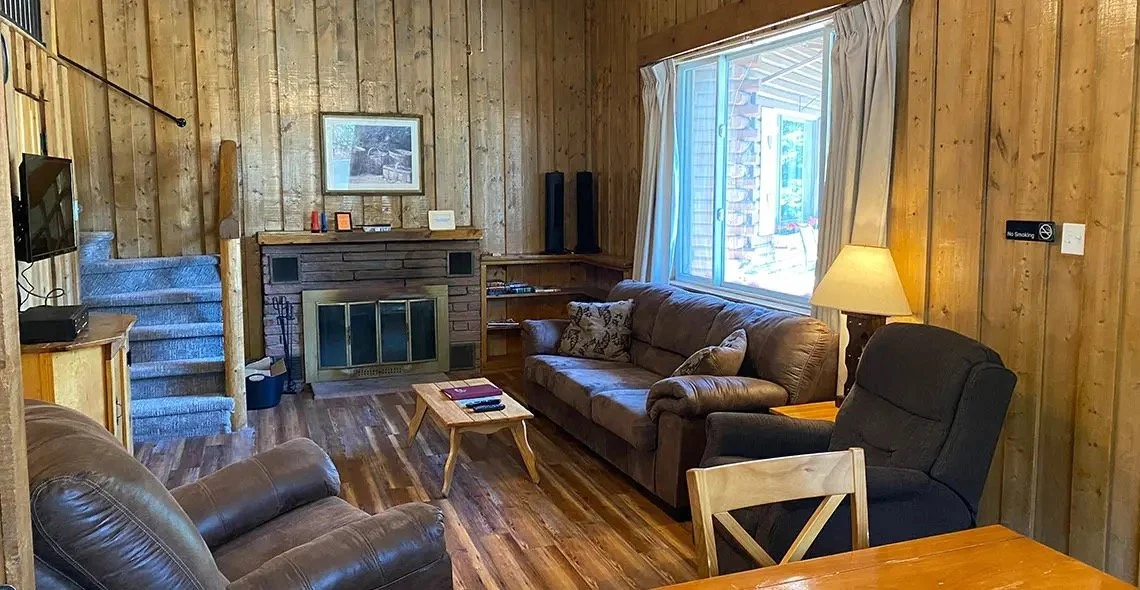 Living room in a cabin with wood-paneled walls, a brick fireplace, a brown leather sofa, a black armchair, a wooden table, a window with white curtains, a lamp, a flat-screen TV, and a staircase with blue carpet.