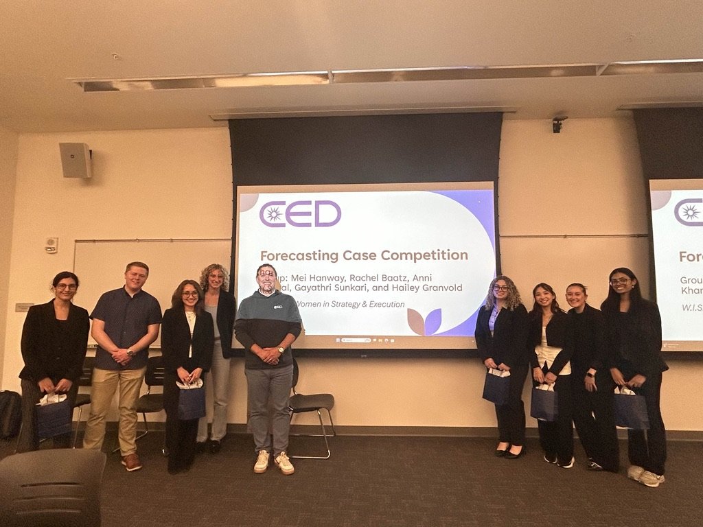 Nine adults standing together next to a projector during the CED Case Competition, dressed in professional attire and posing after delivering their team presentation.