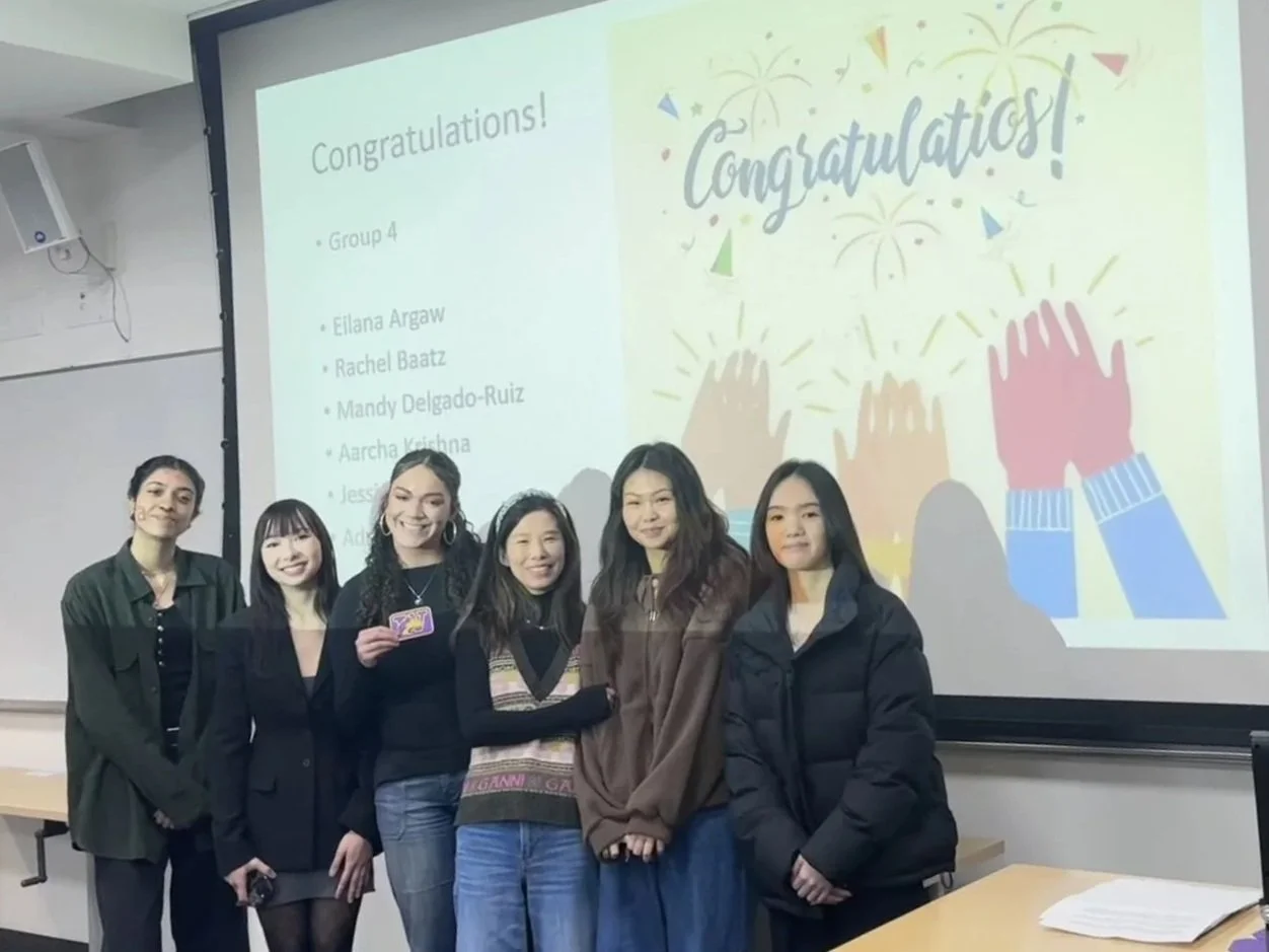 A group of six students standing at the front of a classroom after winning a team competition. Behind them, a projected slide reads ‘Congratulations!’ with celebratory graphics, recognizing Group 4 for their winning presentation.