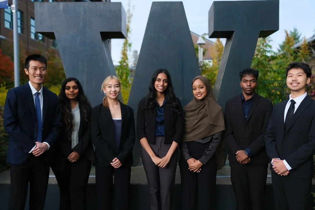 A group of students standing side by side outdoors on the University of Washington Bothell campus, dressed in business and business-casual attire, photographed during a professional headshot session with University of Washington Logo behind.