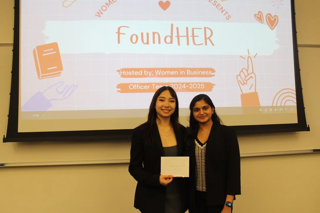 Two students standing in front of a projector during the Women in Business Case Competition, dressed in professional attire and posing after their presentation.