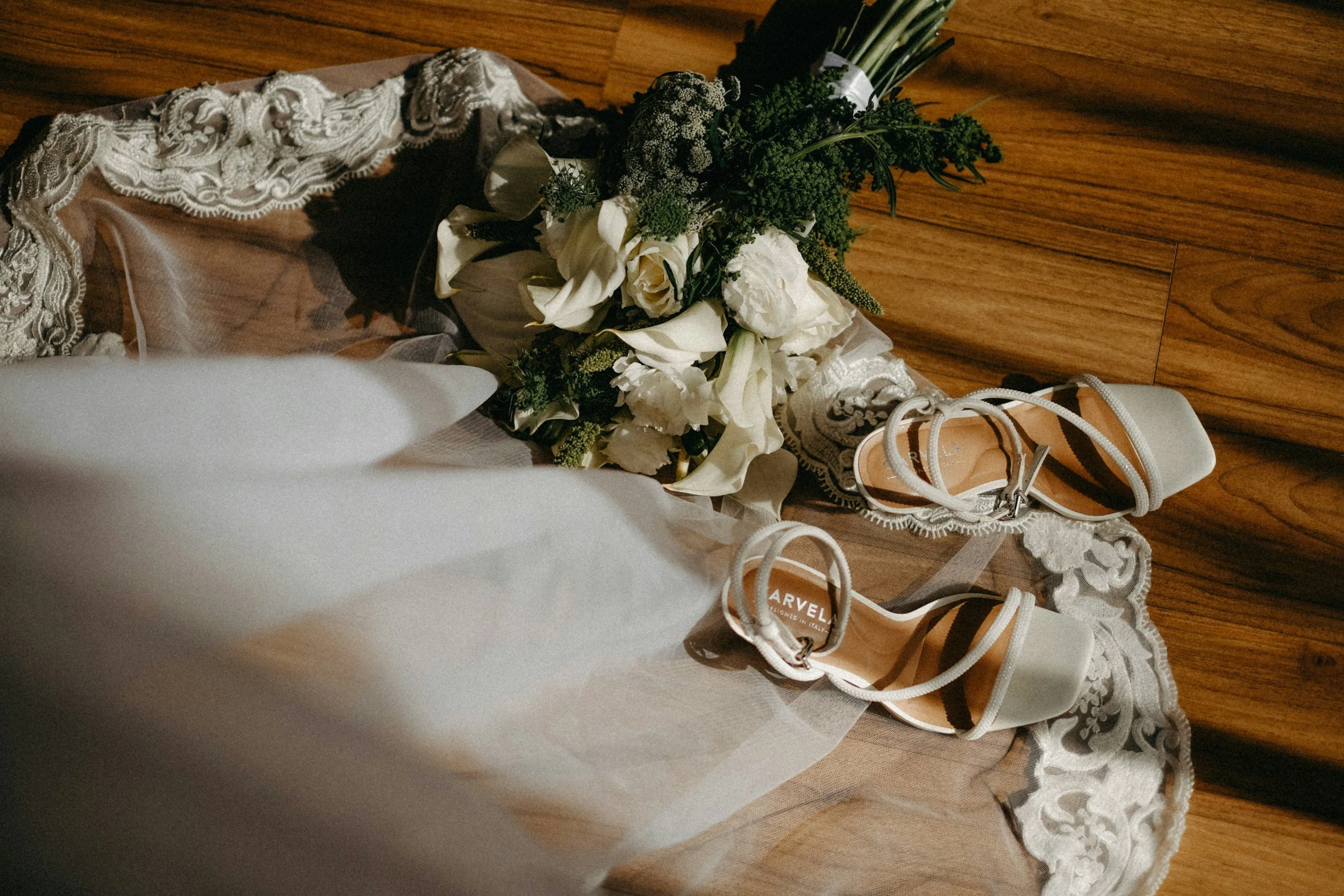A pair of white high-heeled shoes, a bridal bouquet with white roses and greenery, resting on a lace-trimmed cloth on a wooden floor.