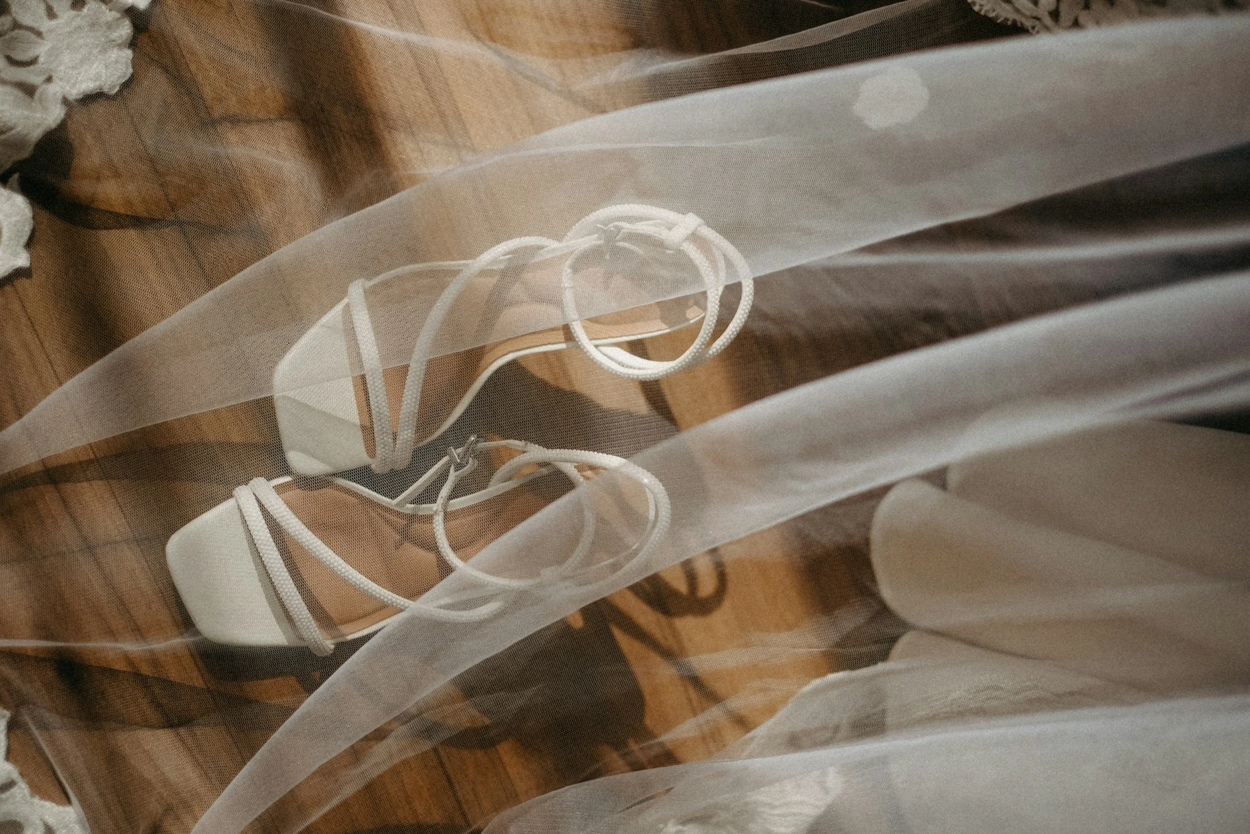White high-heeled sandals with ankle straps resting on a wooden floor underneath a sheer white fabric.