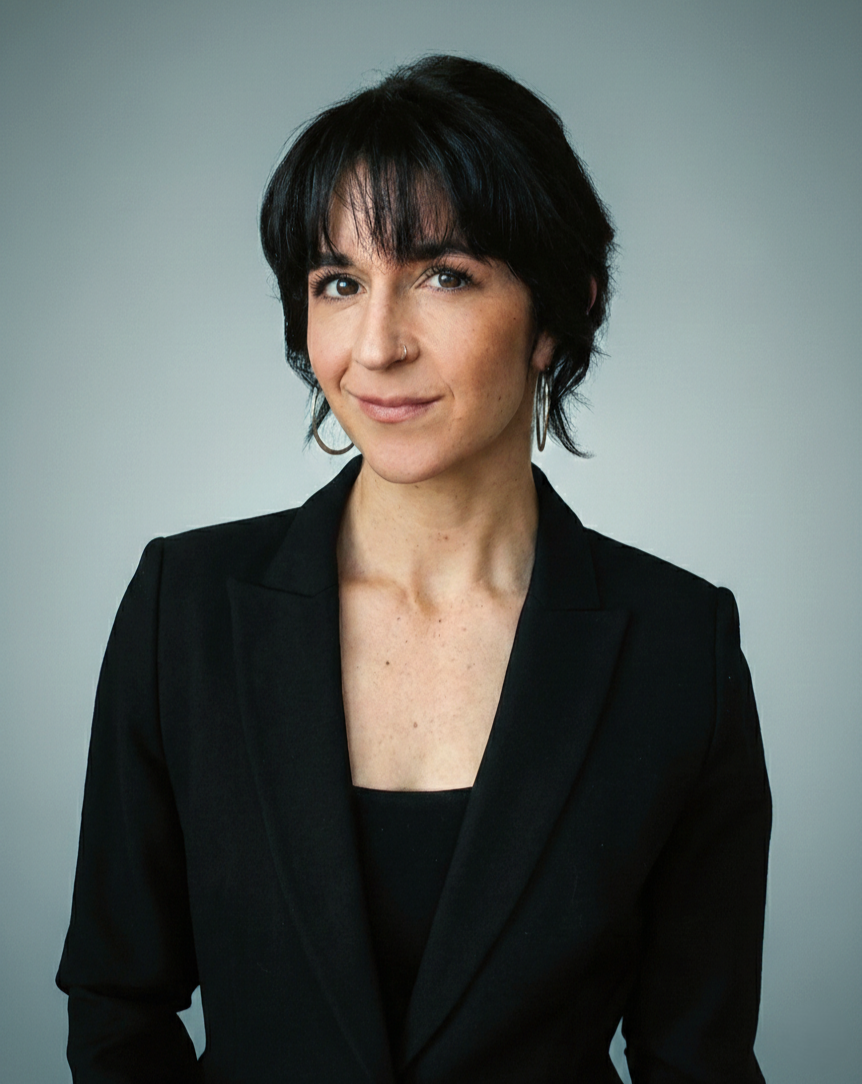 A woman with dark hair, wearing a black blazer and black top, standing against a plain gray background, smiling slightly. Photo portrays the image of President and Principal Consultant of Flora Sustainability Consulting.