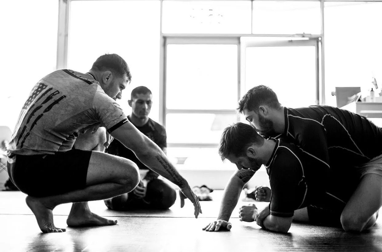 Four men practicing Brazilian Jiu-Jitsu on a mat in a training room, with one instructor demonstrating a technique while the others observe.