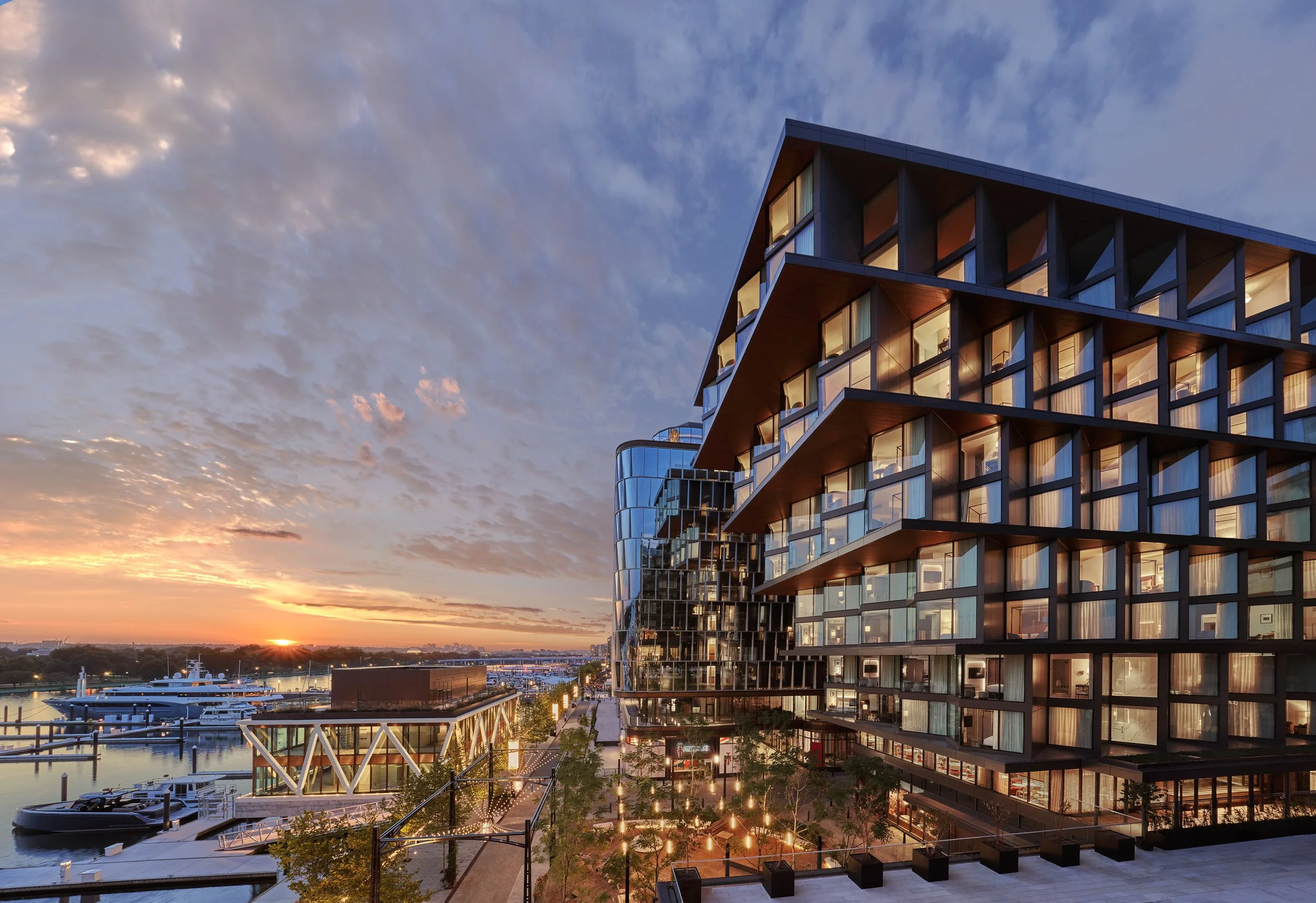 Modern multi-story building with large windows overlooking a waterfront at sunset, with boats docked at a marina.