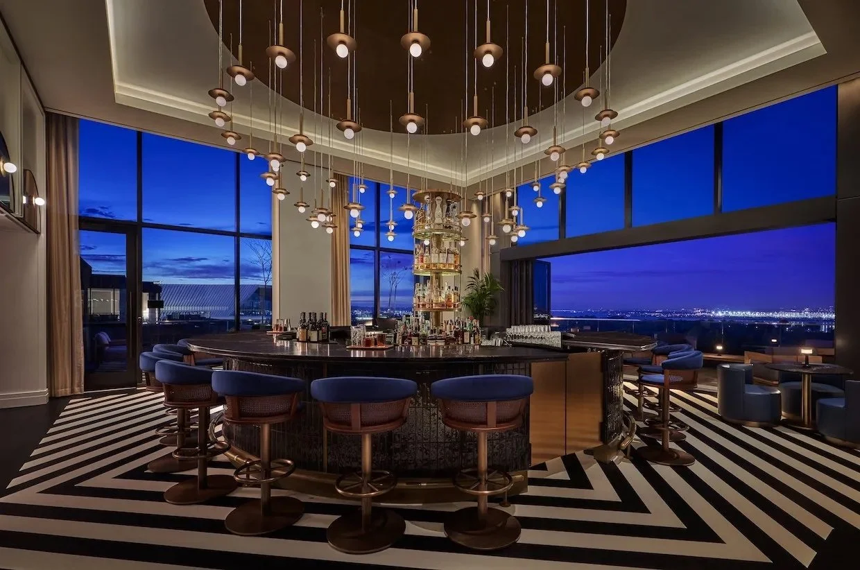 Interior of a modern bar with a circular bar counter, blue upholstered bar stools, large windows showing a cityscape at dusk, and a chandelier with multiple hanging lights.