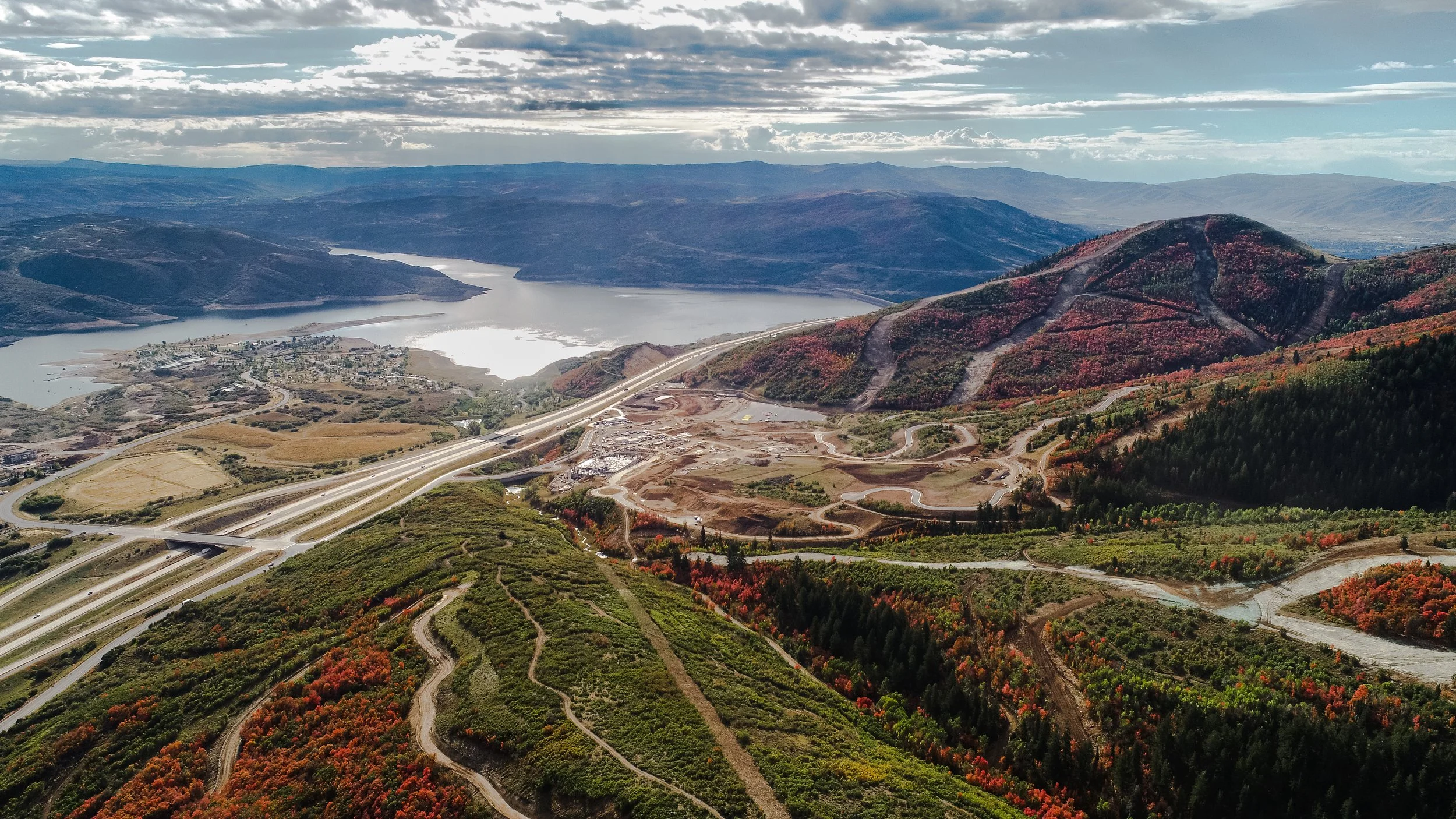 Aerial view of a dam with a large lake behind mountains, winding roads on colorful autumn foliage, and a highway with bridges.