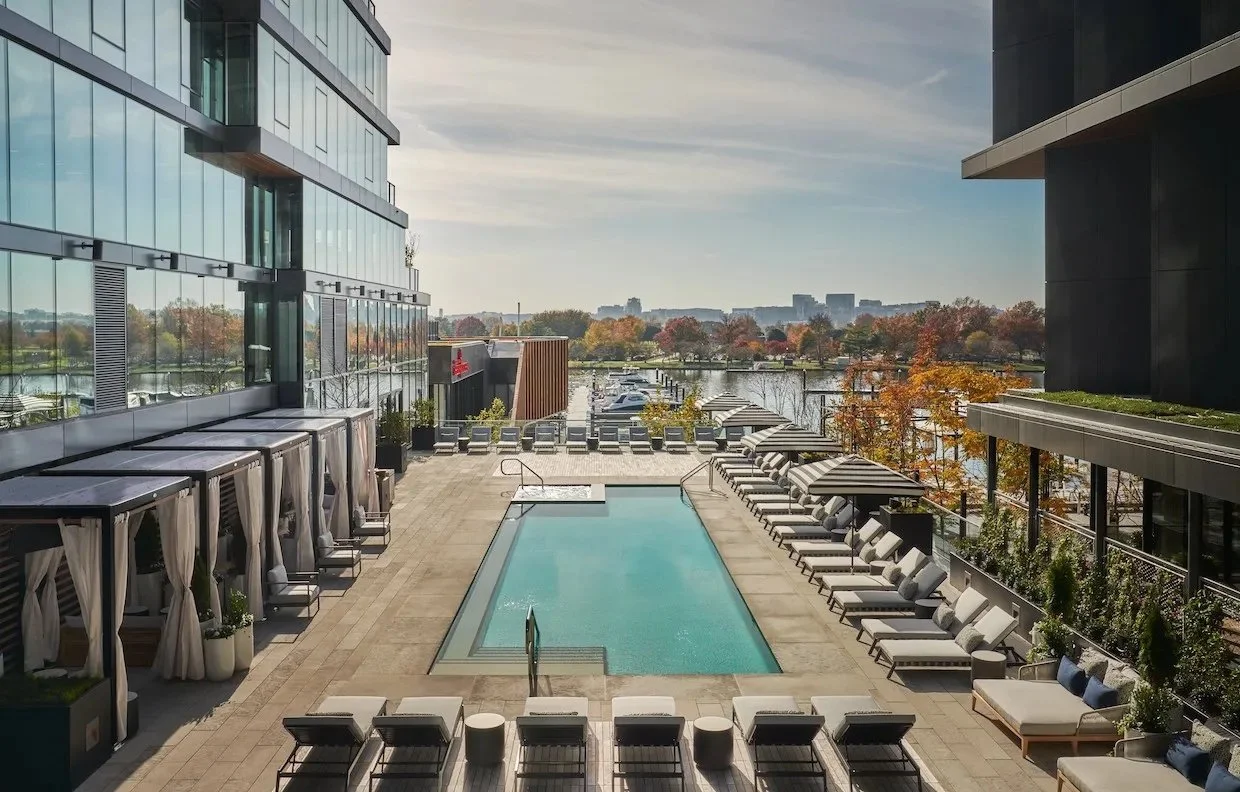 Luxury rooftop pool area with lounge chairs, shaded cabanas, and a view of a river and city skyline in the distance on a clear day.