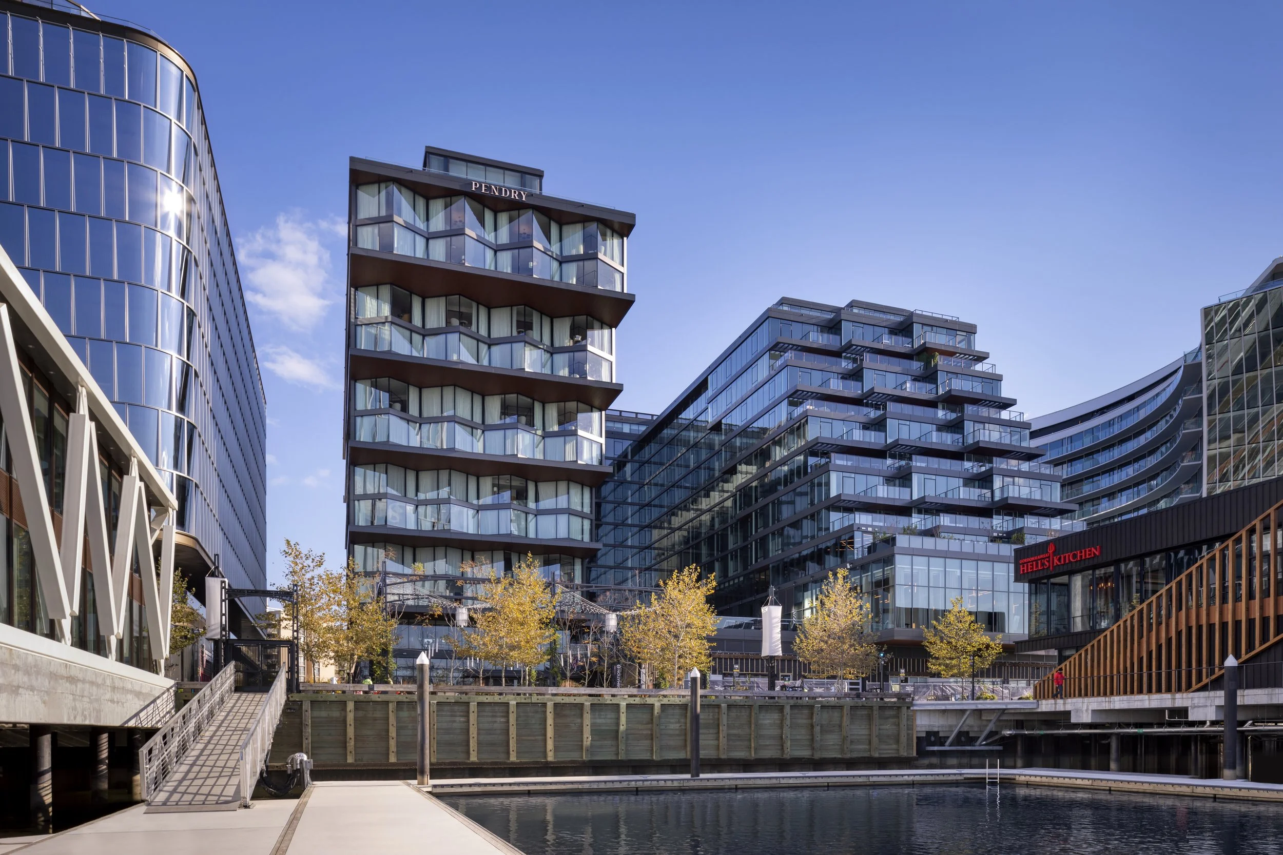 Modern city buildings with reflective glass windows, trees with yellow leaves, and a waterway in the foreground.