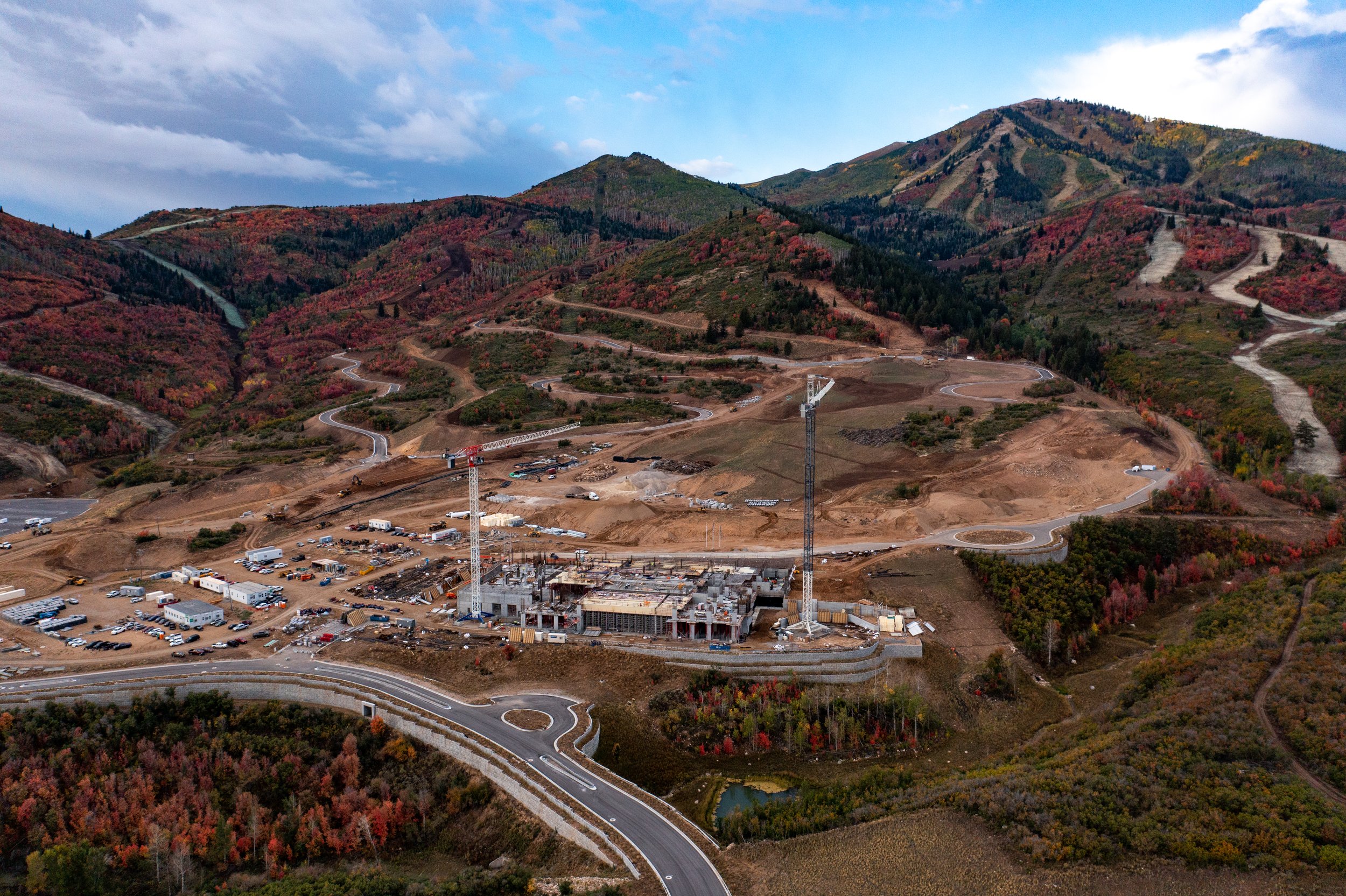 Construction site with cranes and building framework in a mountainous area with winding roads and autumn-colored trees.