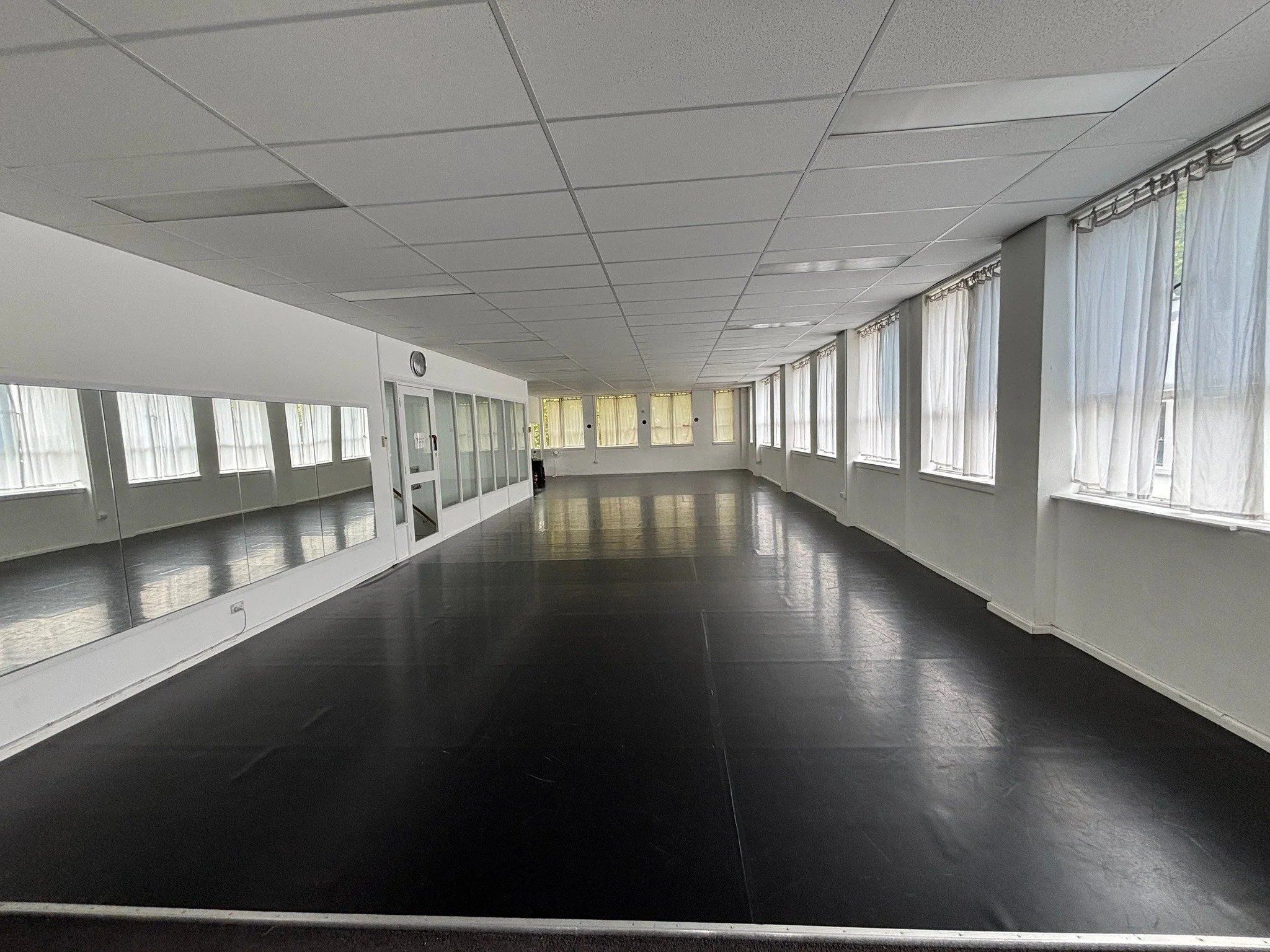 Empty dance or exercise room with large windows covered by curtains, a black floor, white walls, and a mirror along one wall.