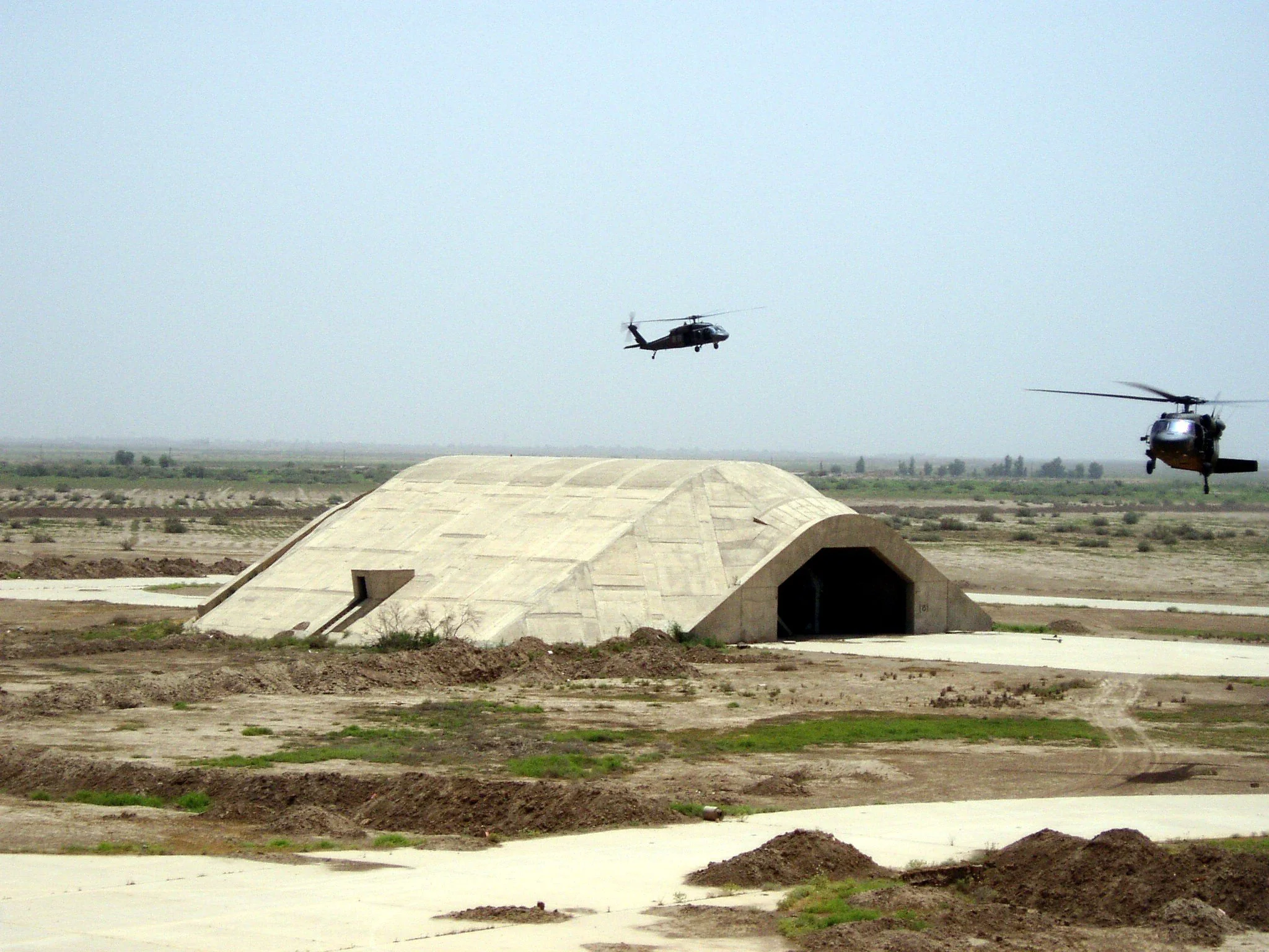 Two military helicopters flying in the sky above a desert with a concrete military bunker.