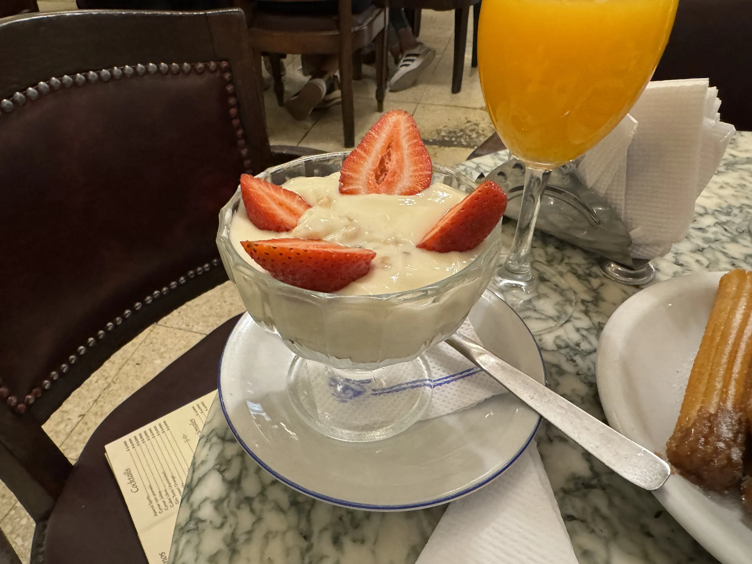 Strawberry shortcake dessert topped with sliced strawberries, a glass of orange beverage, and a piece of caramel churro on a plate on a marble table in a restaurant.