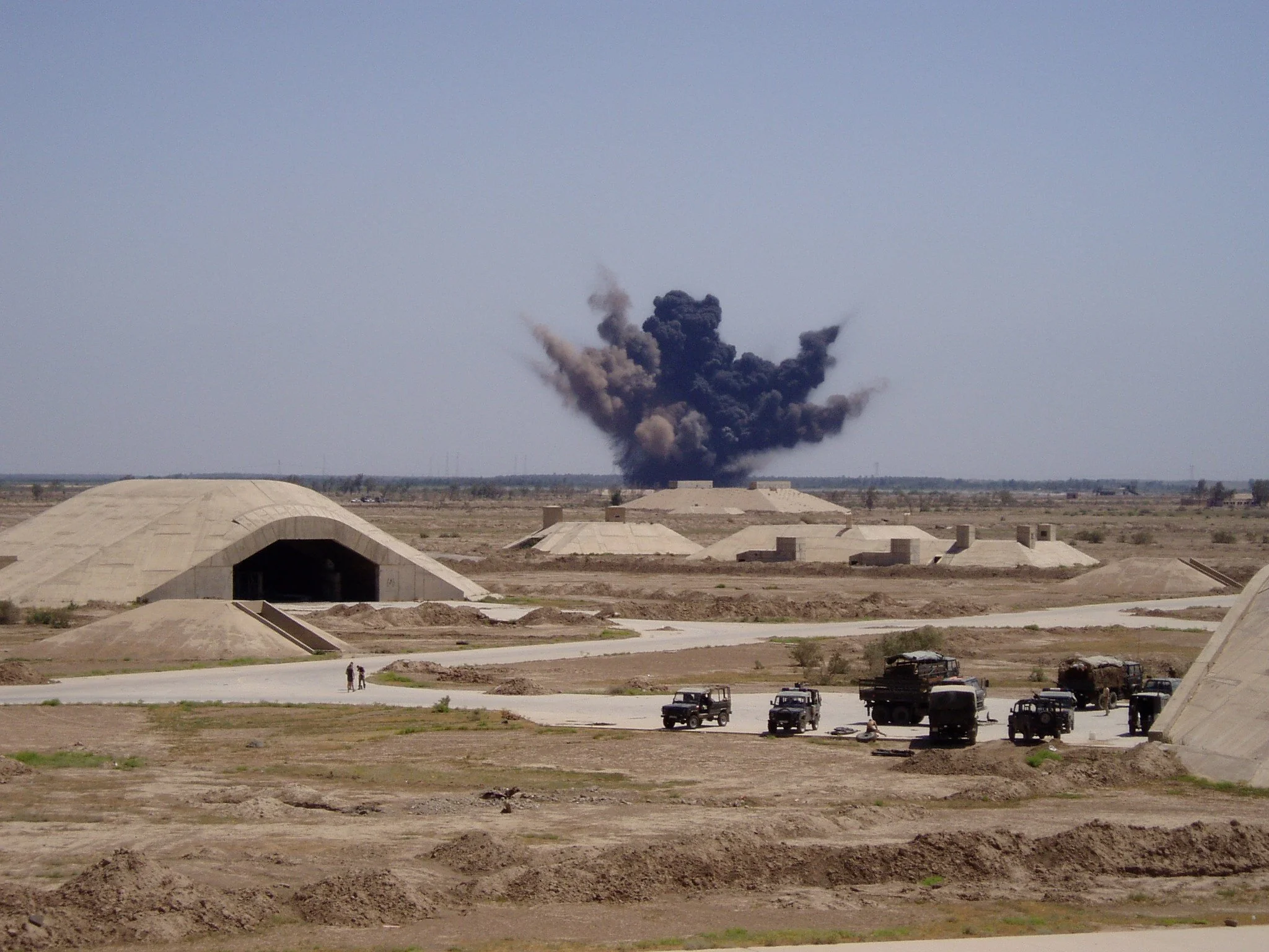 Explosive blast with dark smoke rising from a structure in a desert military base, with military vehicles parked nearby.