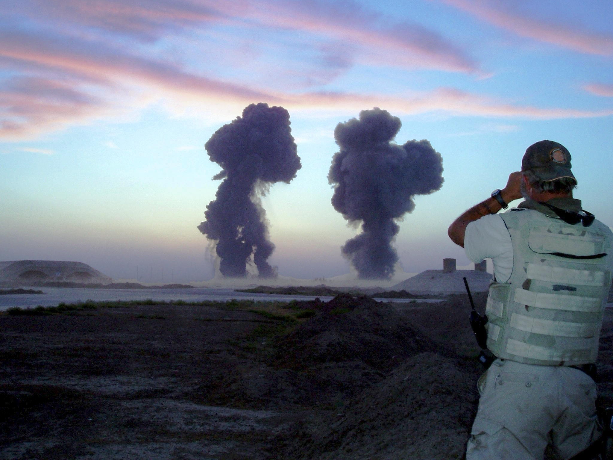 A man in a light-colored tactical vest and a cap is taking a photo or video with a device, possibly a camera, of two large black smoke plumes rising into the sky during sunset or sunrise in a barren landscape.