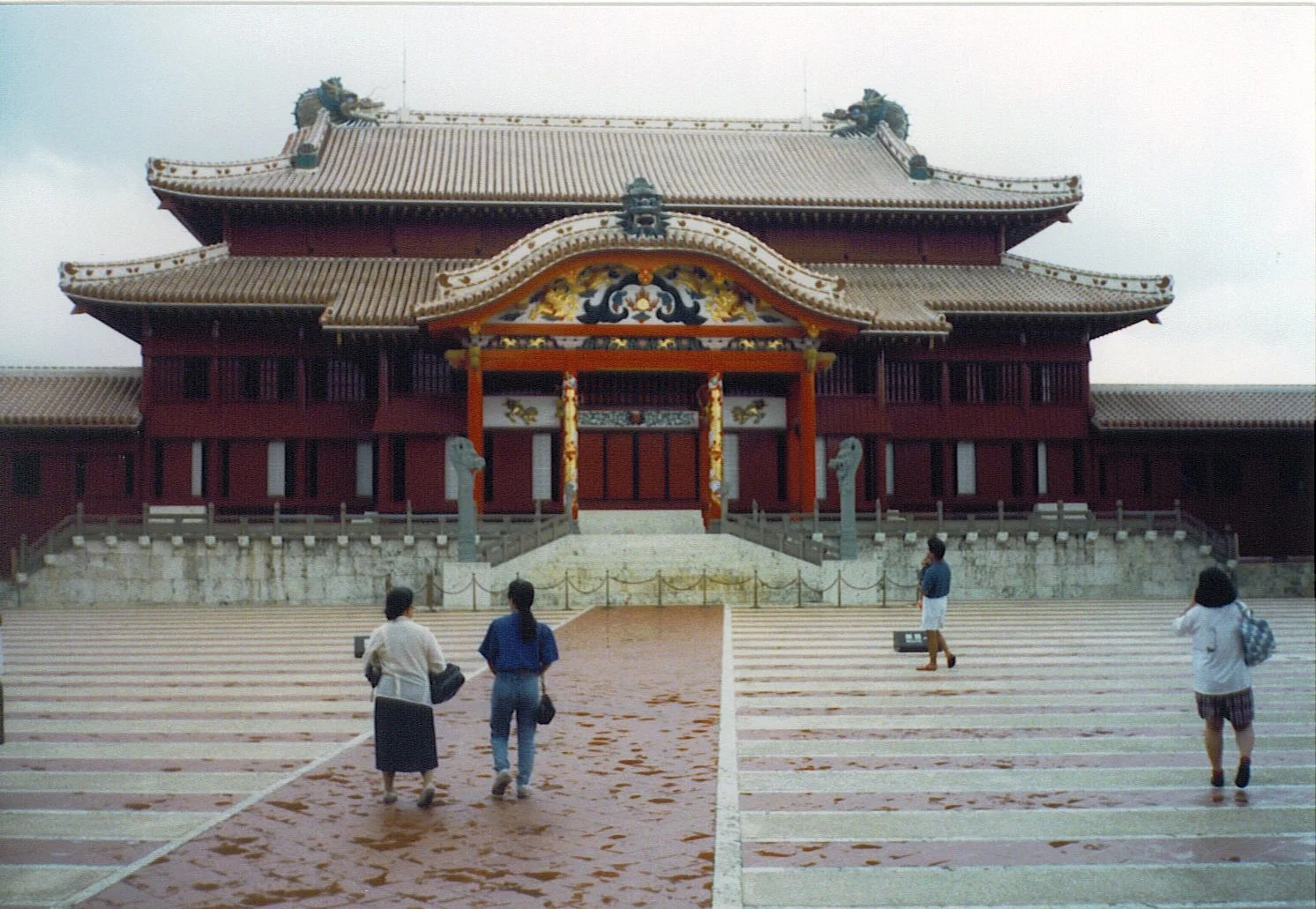 Traditional Japanese temple with ornate roof and decorations, four women walking towards it on a wet, striped stone floor under an overcast sky.