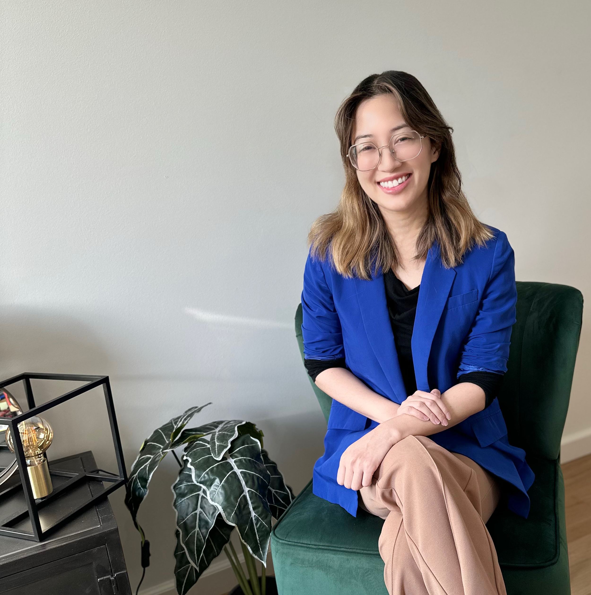 A compassionate looking woman with glasses and an inviting smile, sitting on a green chair, wearing a bright blue blazer and beige pants, with a houseplant and a black side table with decorative items nearby.
