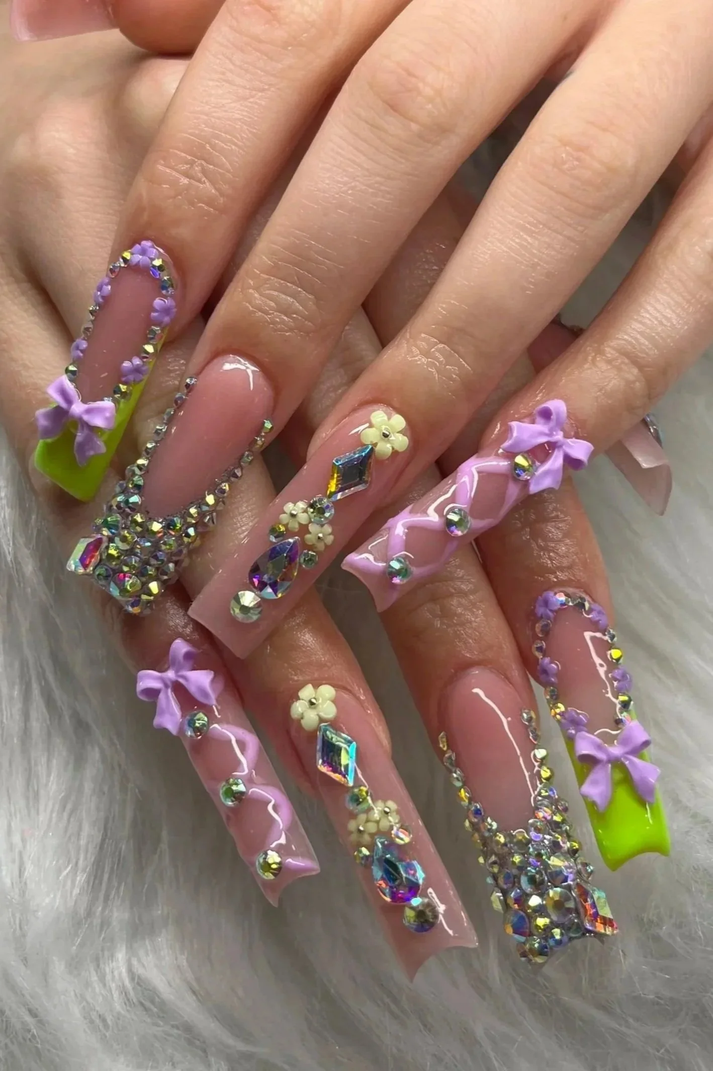 Close-up of a person's hands displaying long, decorated acrylic nails with pastel purple, yellow, and pink designs, adorned with rhinestones, bows, flowers, and 3D embellishments.