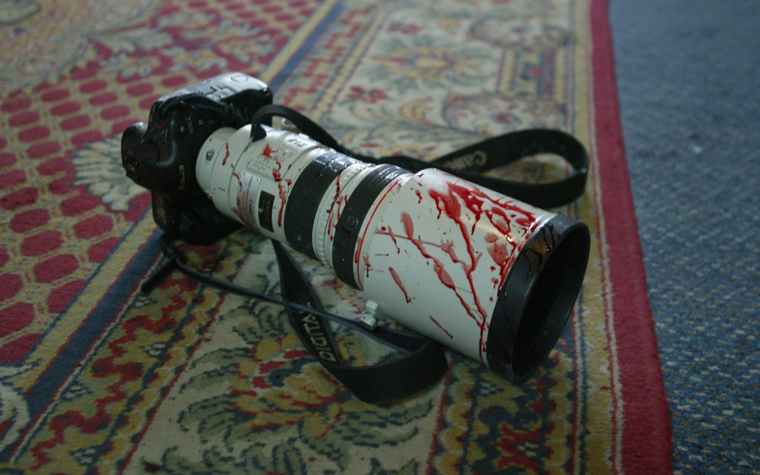 A camera with a long zoom lens lies on a patterned carpet, with red paint splattered all over its white body.