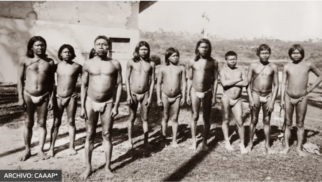 Black and white photo of ten Indigenous children standing outdoors in minimal clothing, with a rural background and simple structures behind them.
