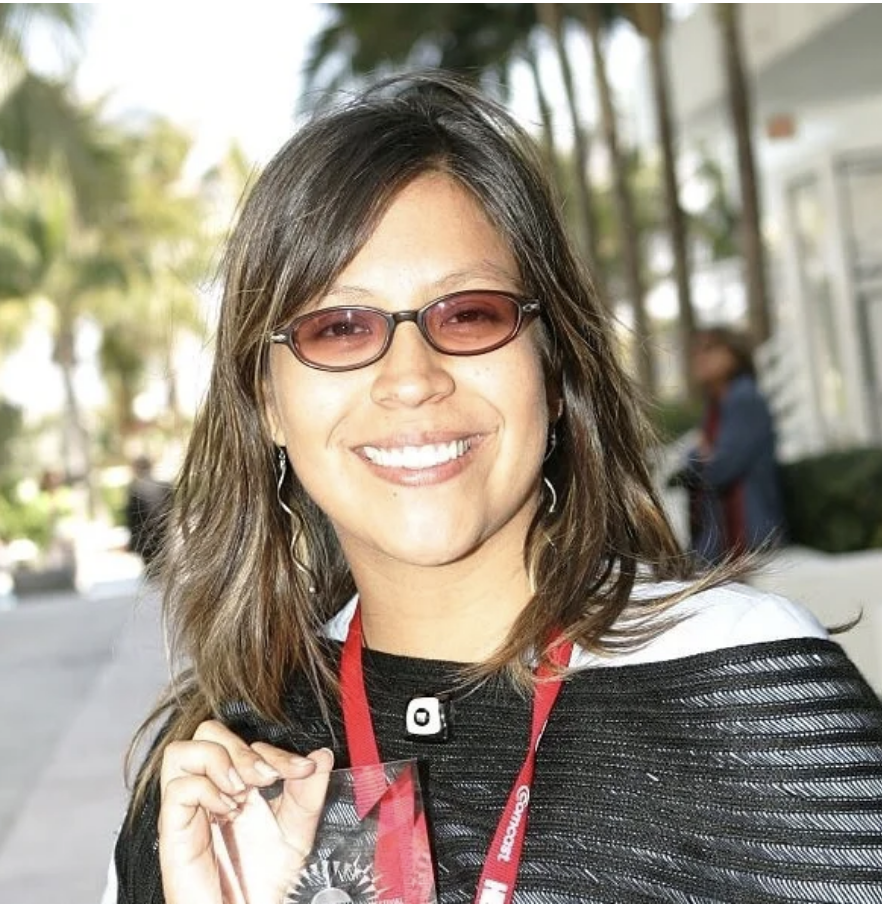 A woman with shoulder-length brown hair wearing sunglasses, smiling, holding a clear glass award, and wearing a black and white top with red lanyard outdoors.