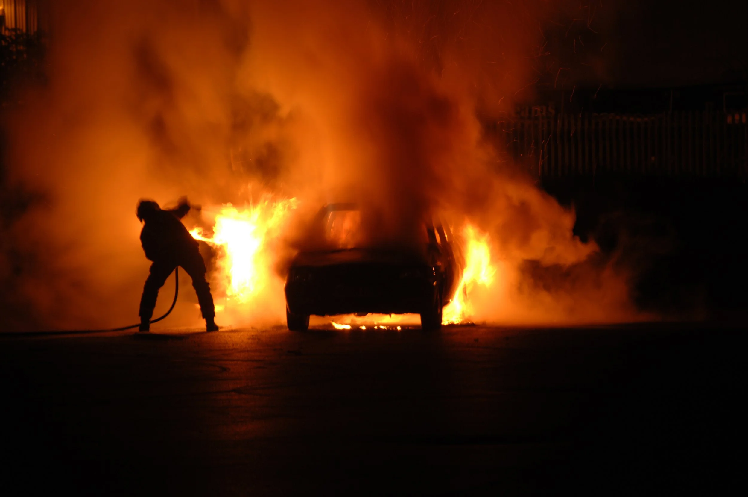 A person fighting a fire in a car with flames and thick smoke at night.