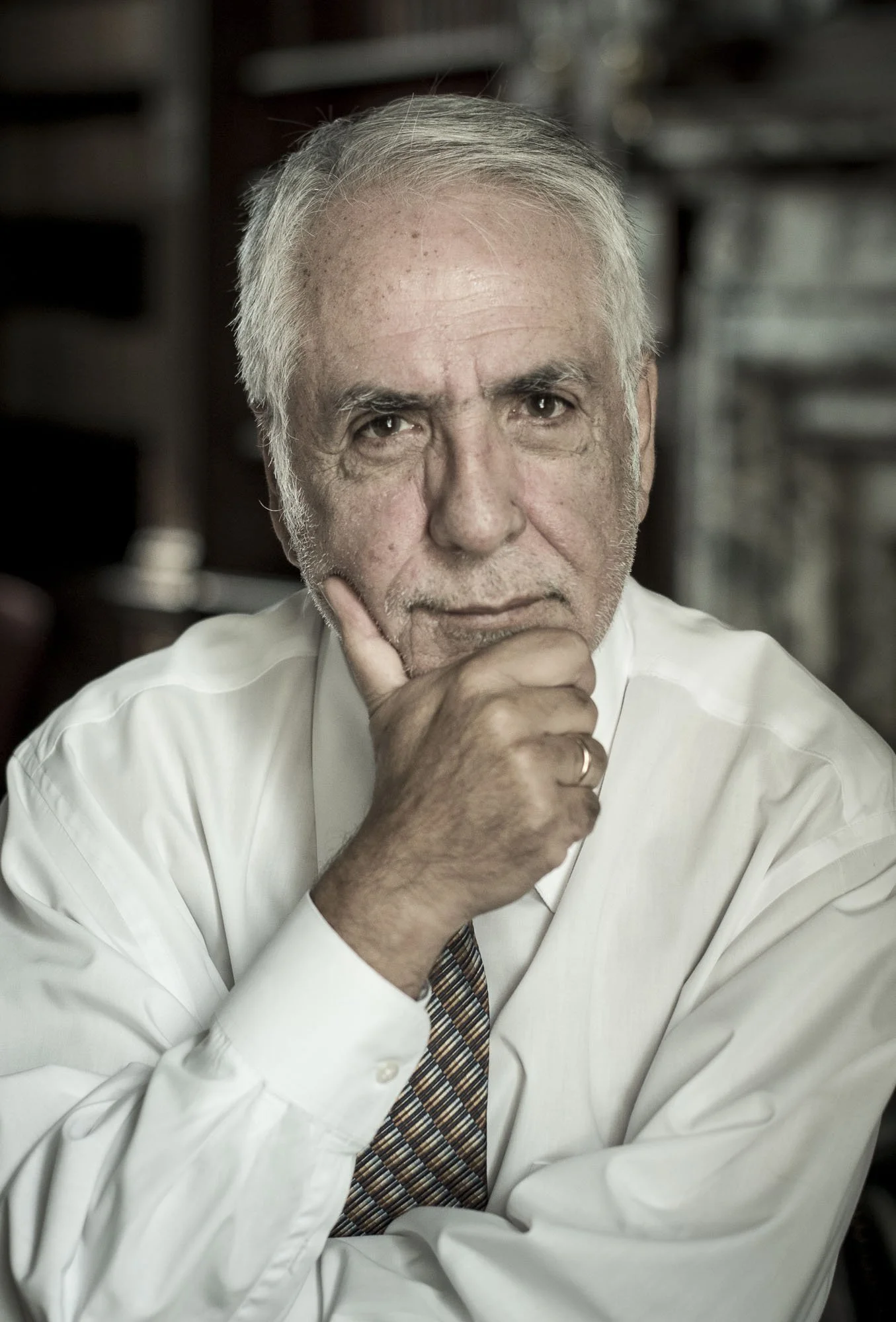 A senior man with white hair and a serious expression, resting his chin on his hand while looking at the camera in a professional setting.