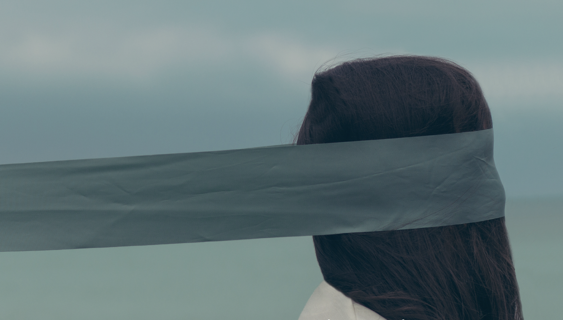 A person with long dark hair facing sideways, with a grayish cloth or tape covering their eyes, against a cloudy sky background.