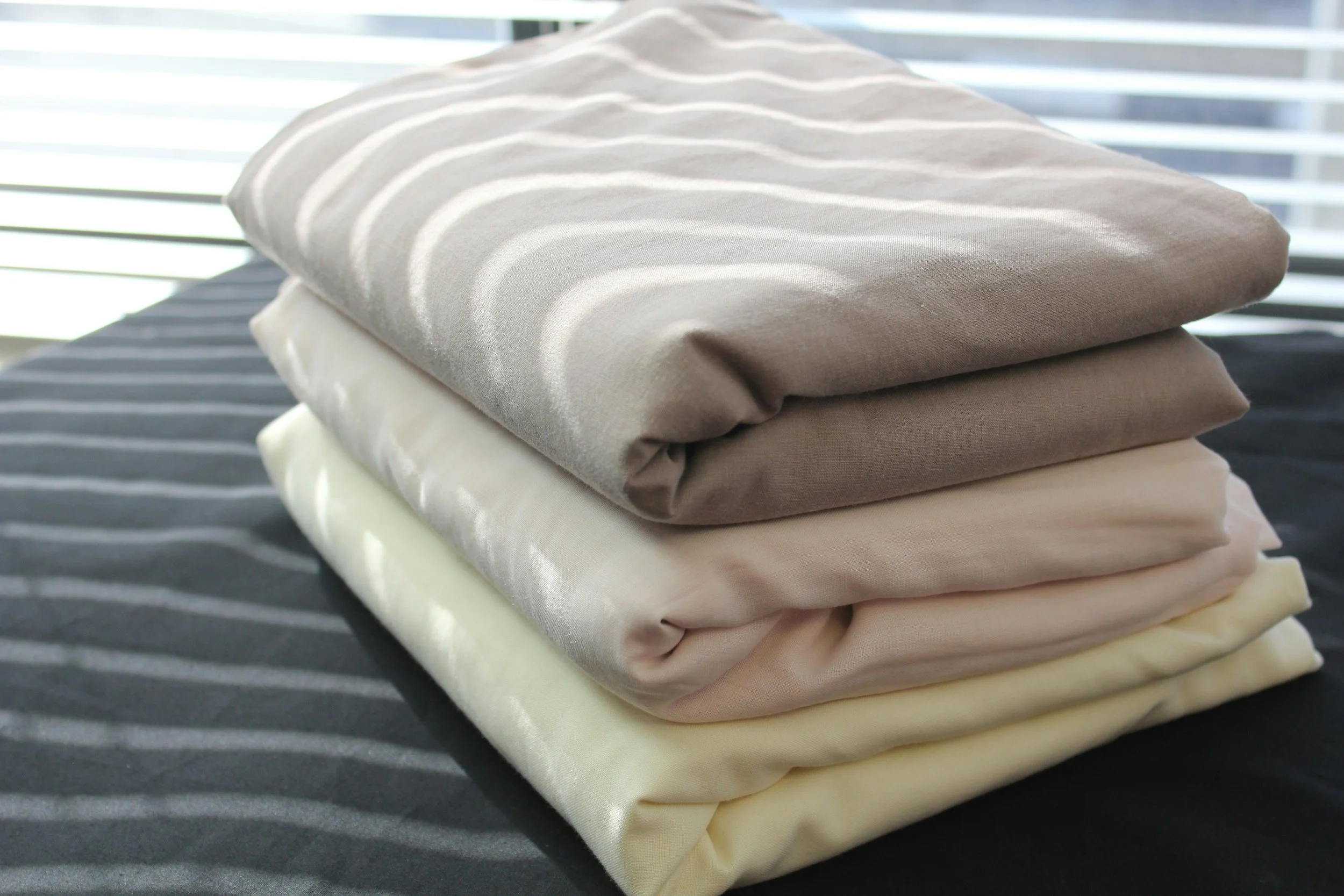 A neatly folded stack of five bedsheets in shades of beige, cream, and taupe, placed on a dark surface near window blinds.