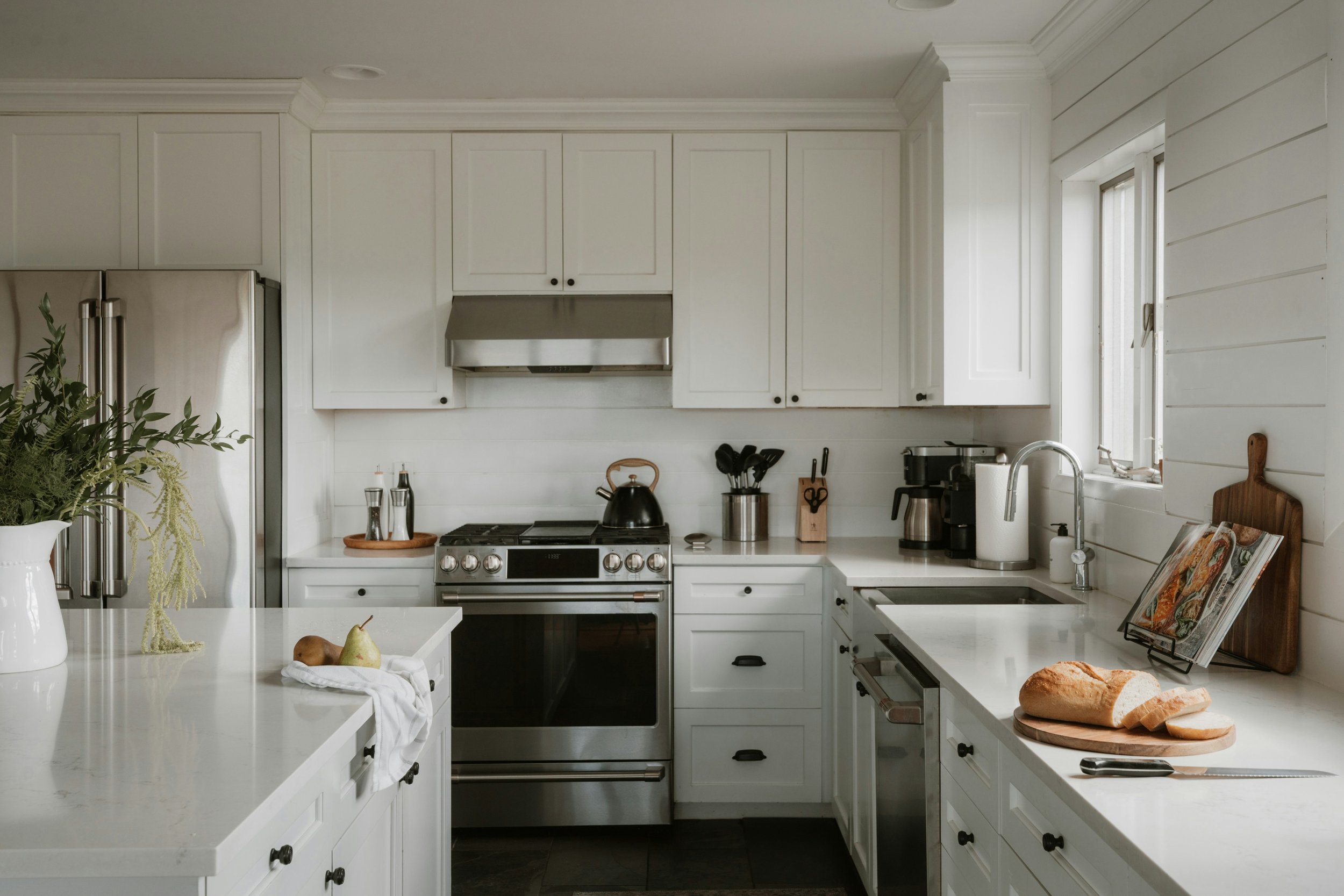 Clean white kitchen with stainless steel appliances, a loaf of bread on a cutting board, and cooking utensils on the counter.