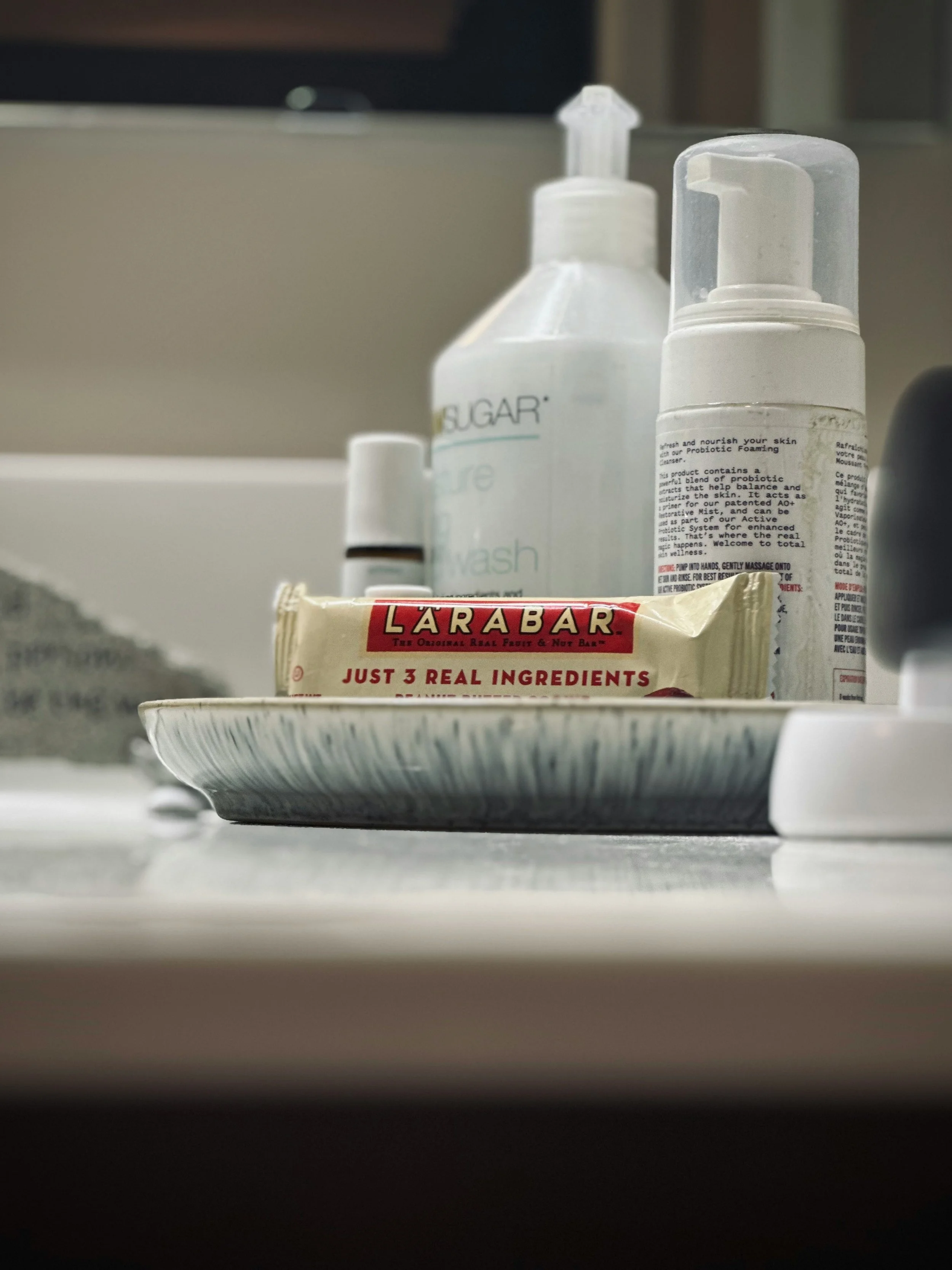 A close-up of a dish with a LÄRABAR protein bar on top, alongside various personal care bottles in the background, including a pump soap dispenser, a spray bottle, and a small container.