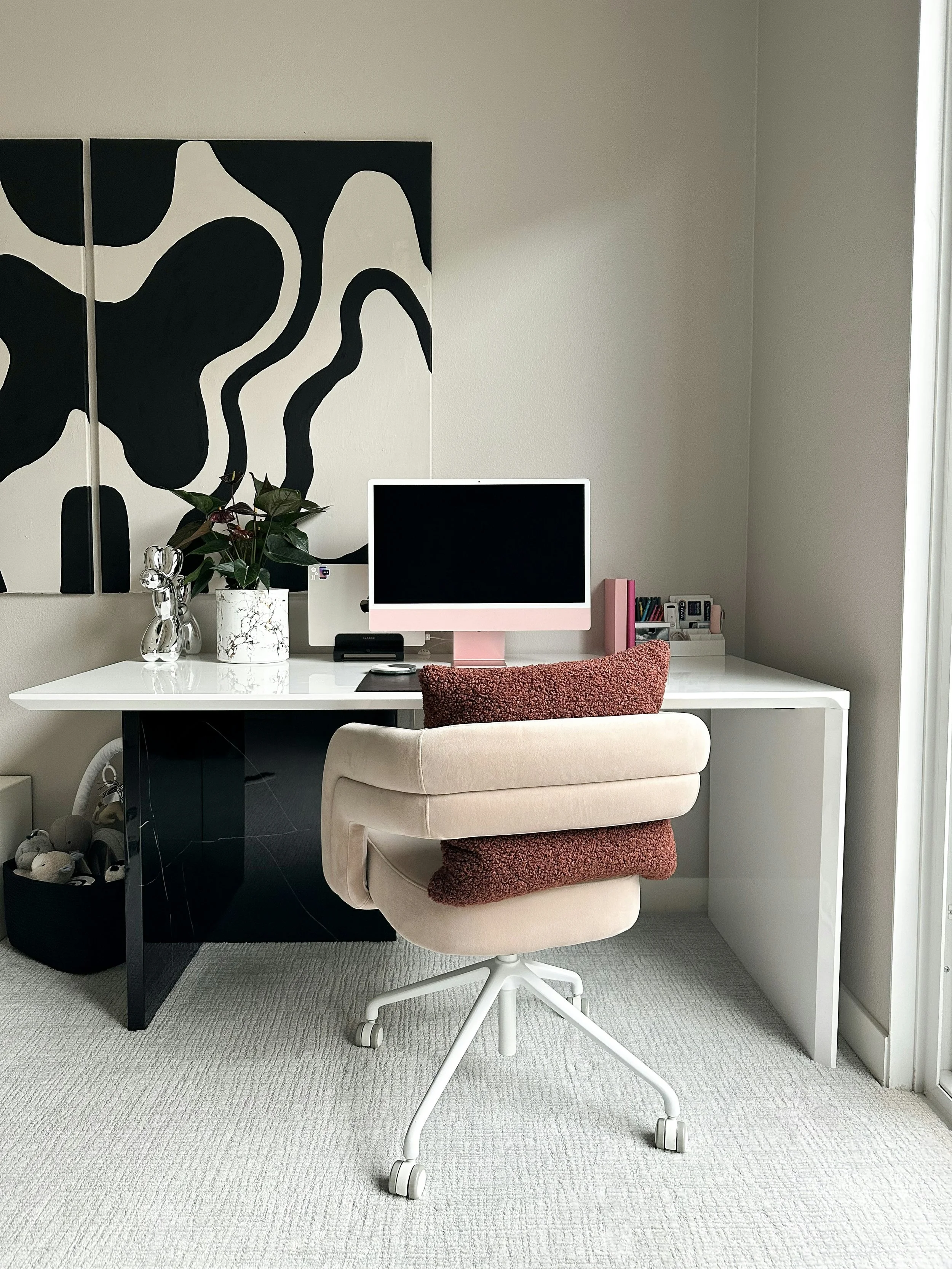 A home office with a white desk, a pink computer monitor, a beige swivel chair with red cushions, a black and white abstract painting on the wall, a potted plant, and various office supplies on the desk.