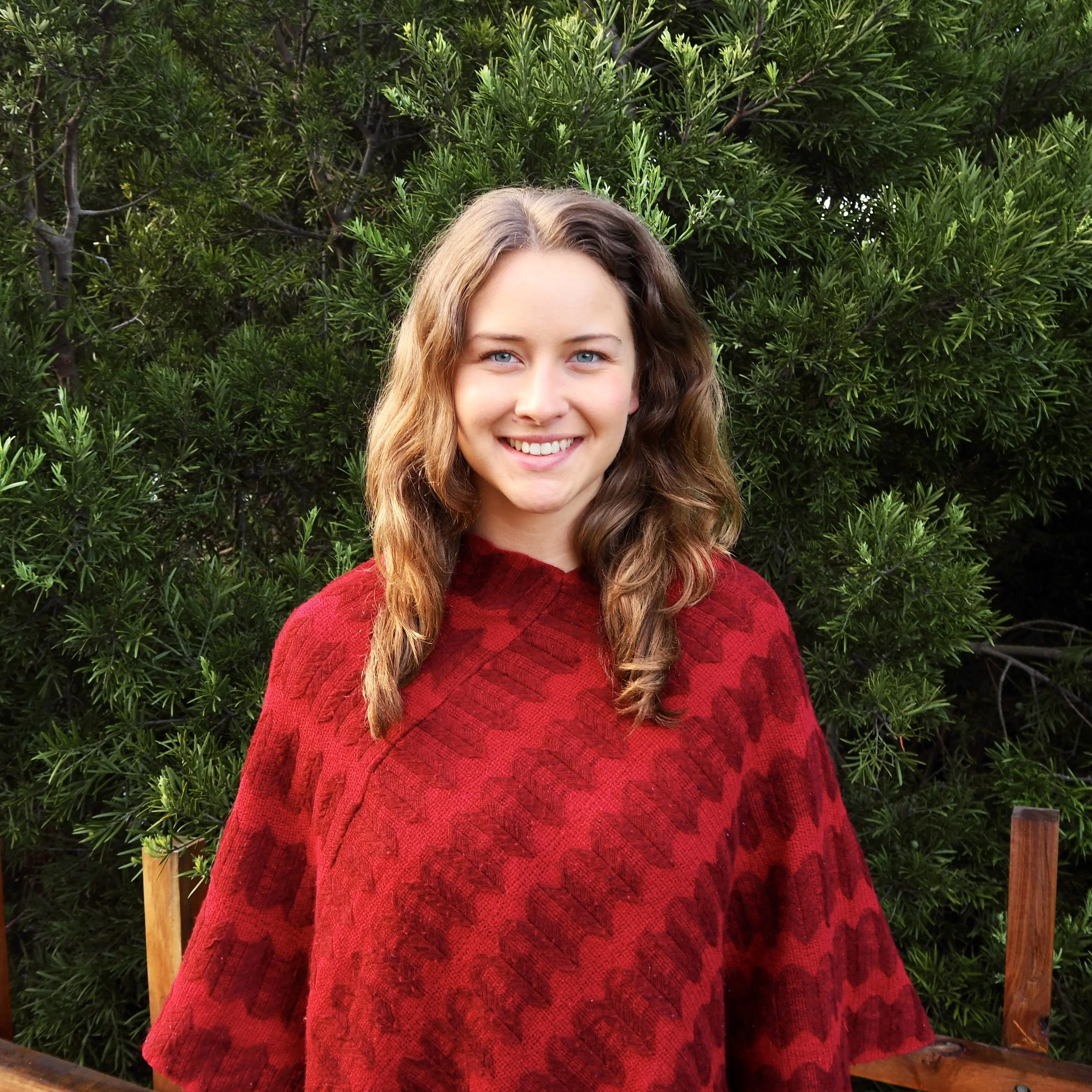 Smiling young woman with wavy brown hair wearing a red shawl standing outdoors in front of greenery.