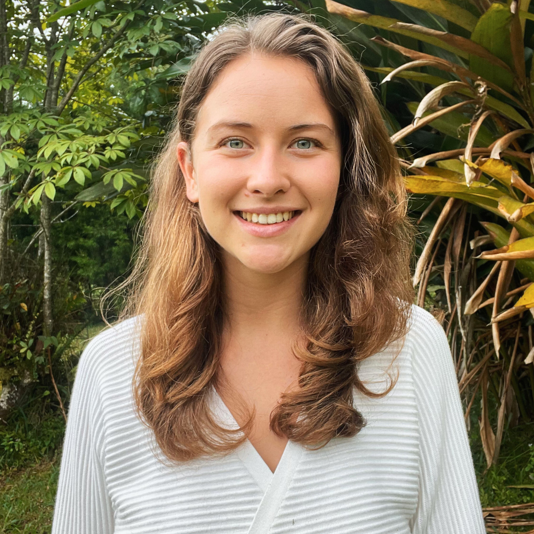 A young woman with long, wavy brown hair and blue eyes smiling outdoors, surrounded by green foliage.