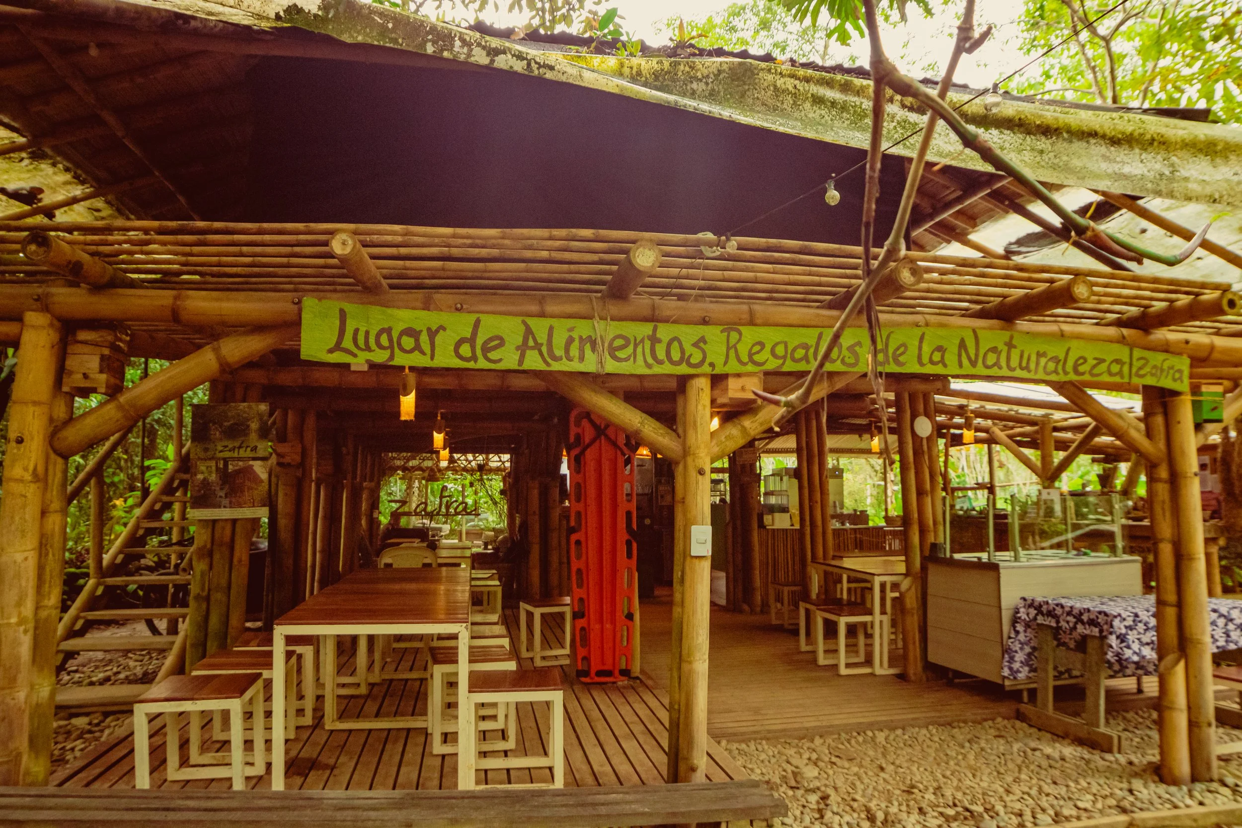 A rustic outdoor market stall made of bamboo with a sign in Spanish that reads "Lugar de Alimentos, Regalos de la Naturaleza". The stall has tables and chairs inside, with a roof made of bamboo and a natural setting with trees.