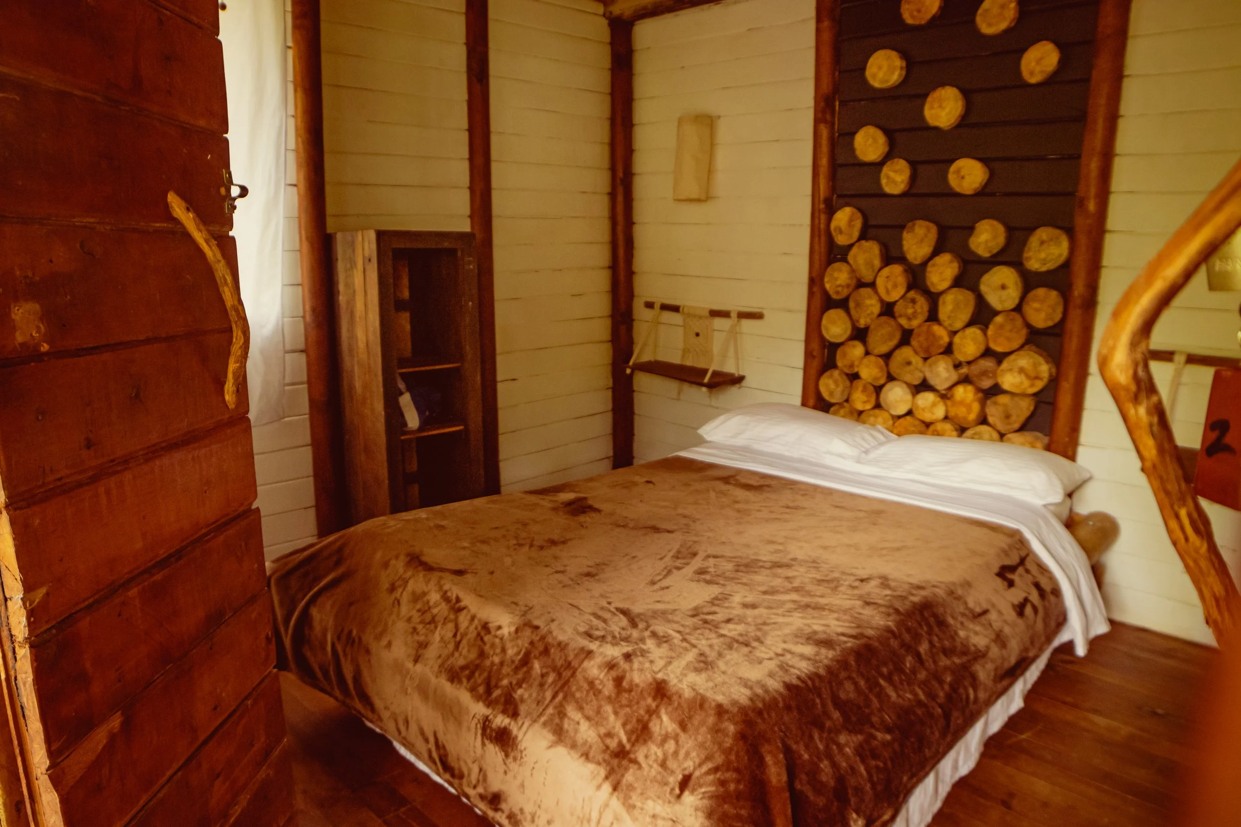 A cozy bedroom with wooden walls, a bed with white pillows and a brown blanket, a small bookshelf, and a decorative wall feature made of stacked logs.