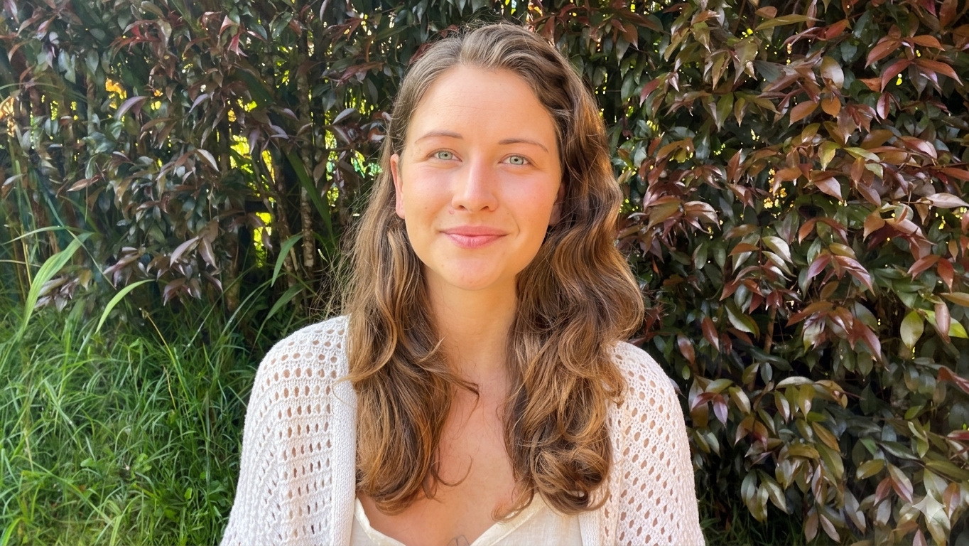 A woman with long, curly brown hair and light skin is smiling outdoors in front of green and purple-leafed bushes, wearing a white top and a white, open-knit cardigan.