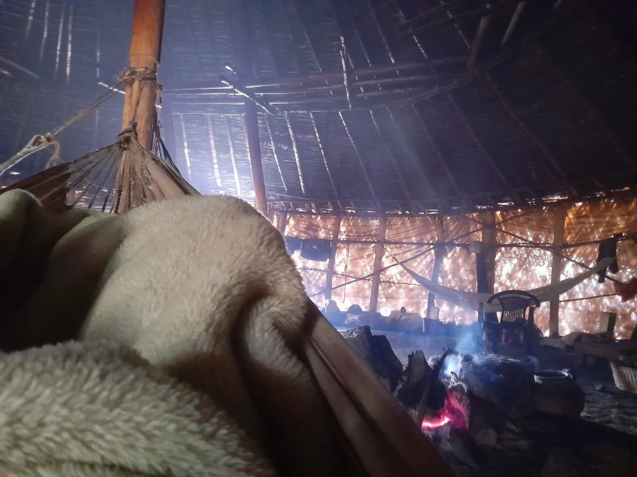 Inside a thatched hut with a coat and a hammock hanging, fire burning on a stone platform with smoke rising, and daylight filtering through the gaps in the roof.