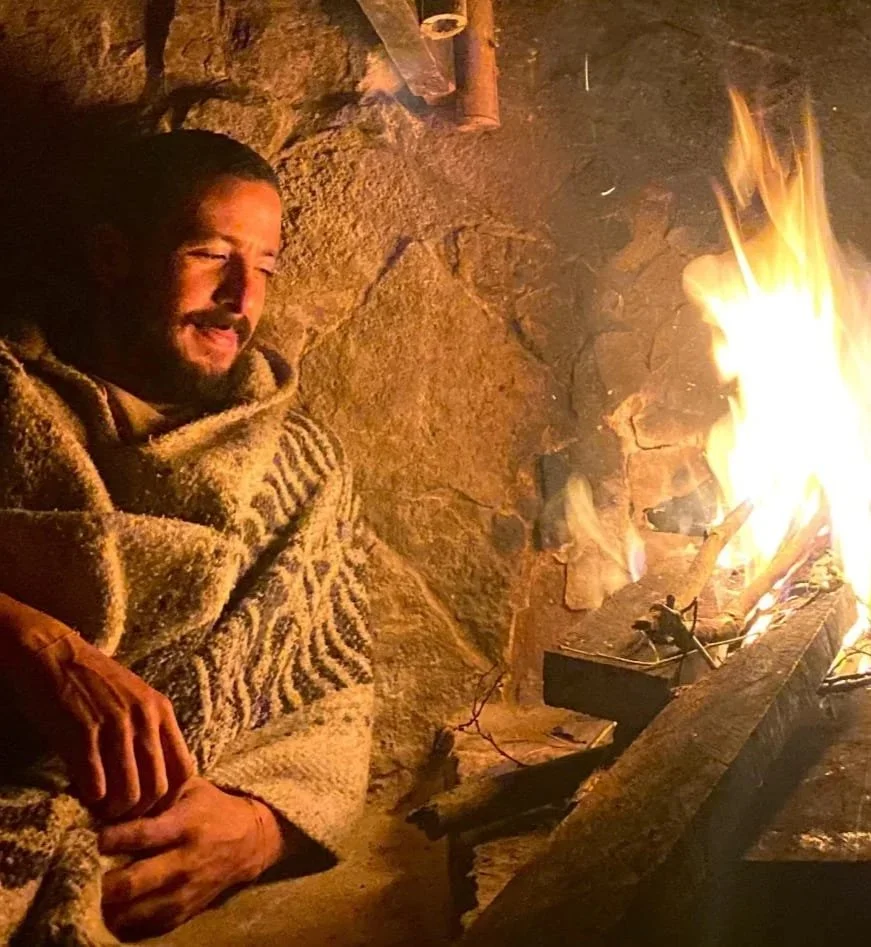 A person with a beard and long hair wrapped in a blanket, sitting near a campfire inside a stone-walled shelter.
