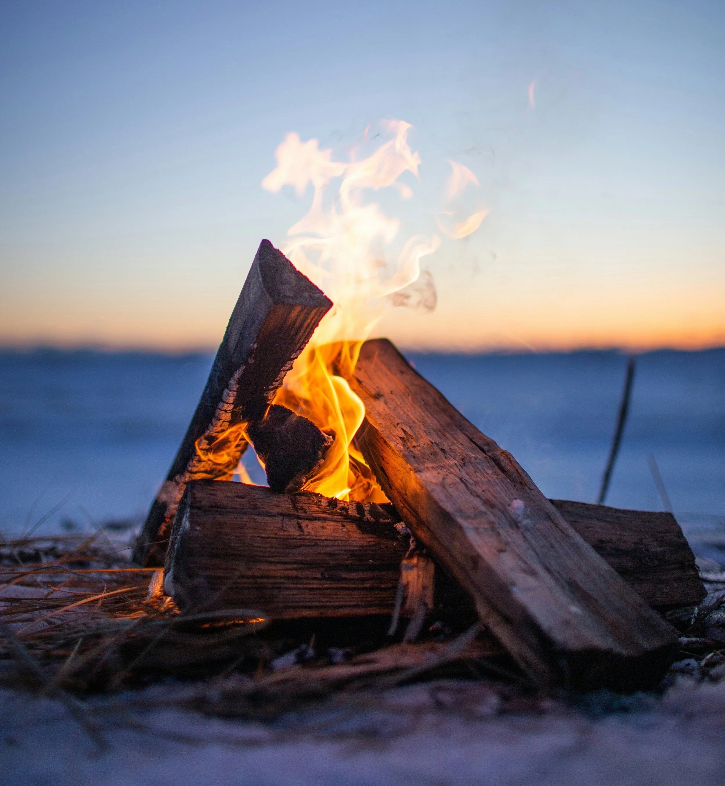A campfire burning with several logs, outdoors at sunset, with a blue sky and distant horizon in the background.