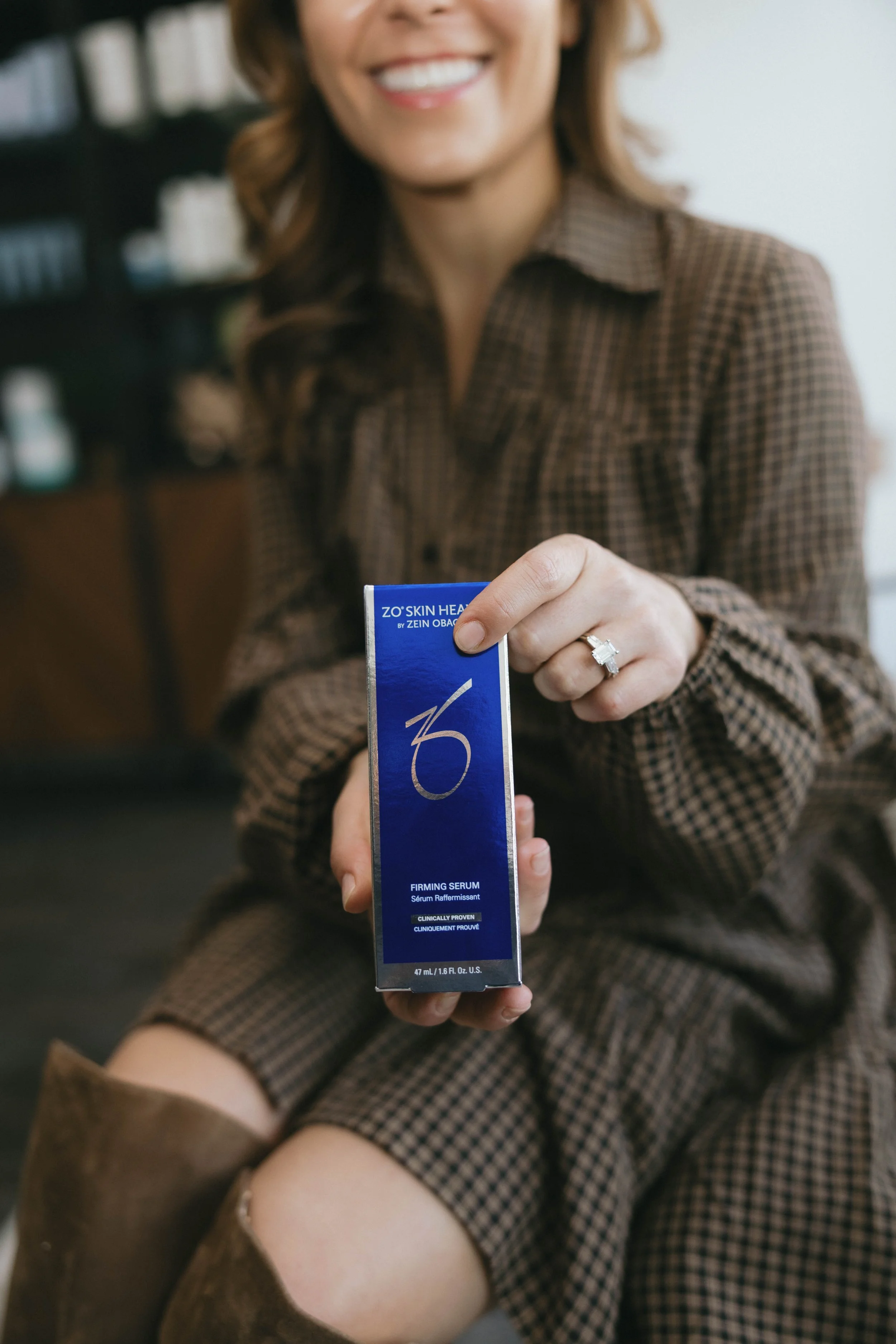 A woman smiling and showing a box of skincare product labeled 'ZO Skin Health' with her left hand, wearing a checkered brown shirt and a wedding ring, sitting indoors.