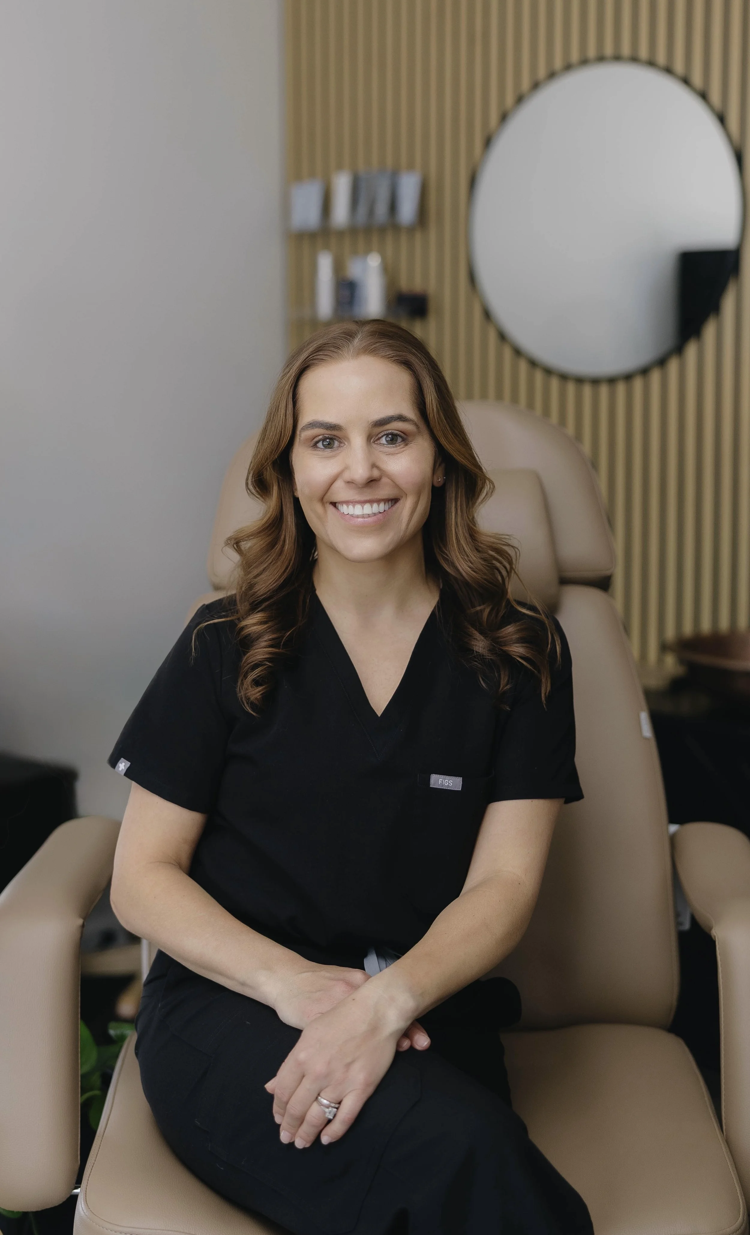 A woman with brown hair styled in loose waves sitting on a beige chair inside a modern office or clinic space, smiling at the camera, wearing a black medical uniform.