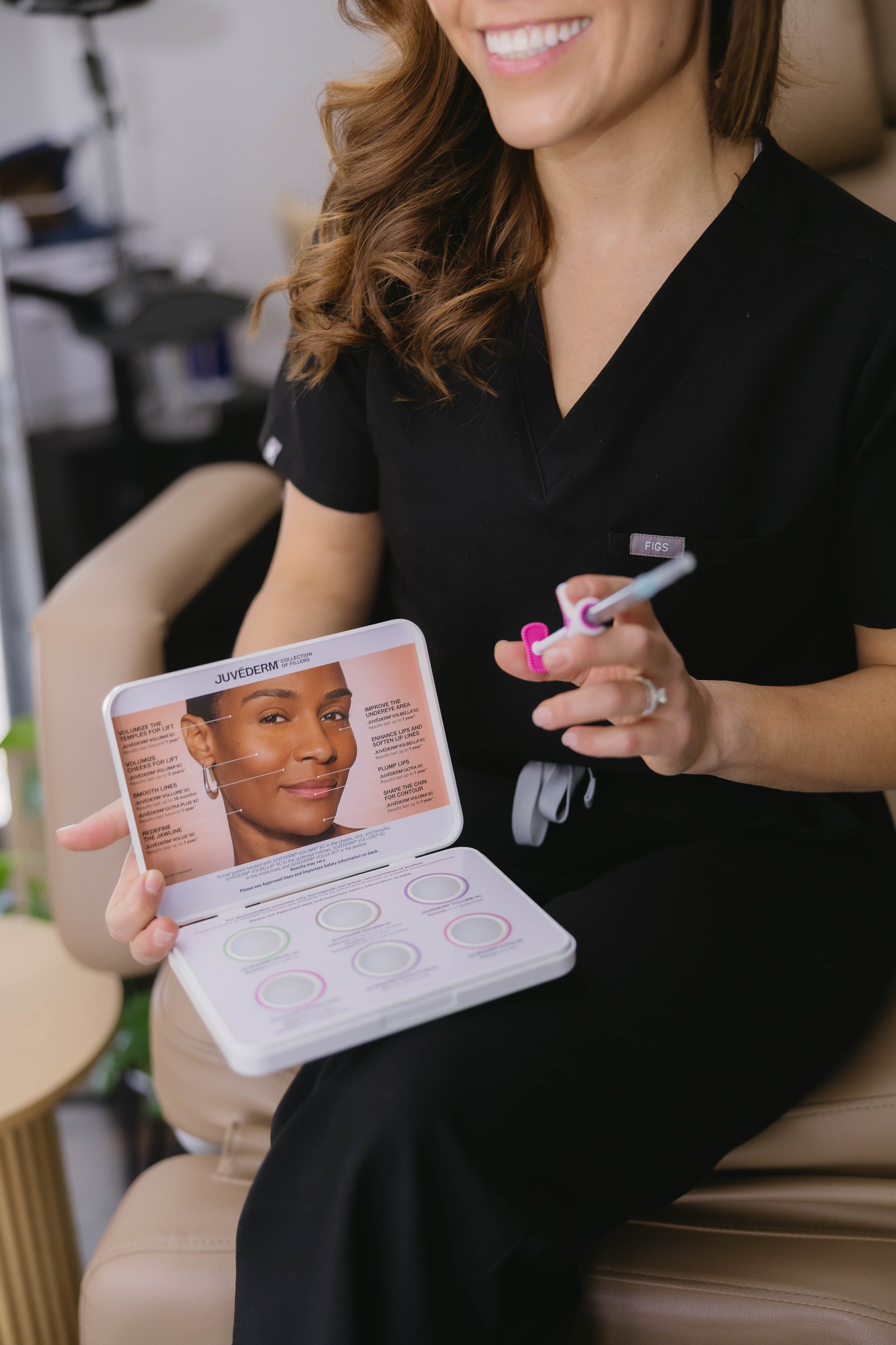 A woman in black scrubs sitting on a beige chair, smiling. She holds a small open box with skincare product descriptions and a color-matching tool, and a tool with pink components.