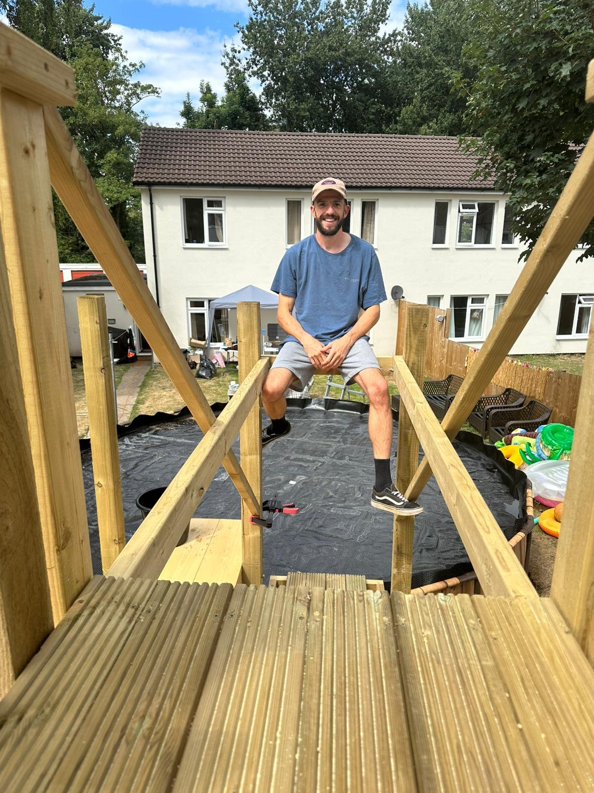 Caro sitting on the wooden frame of a deck under construction in a backyard, smiling with a house in the background.