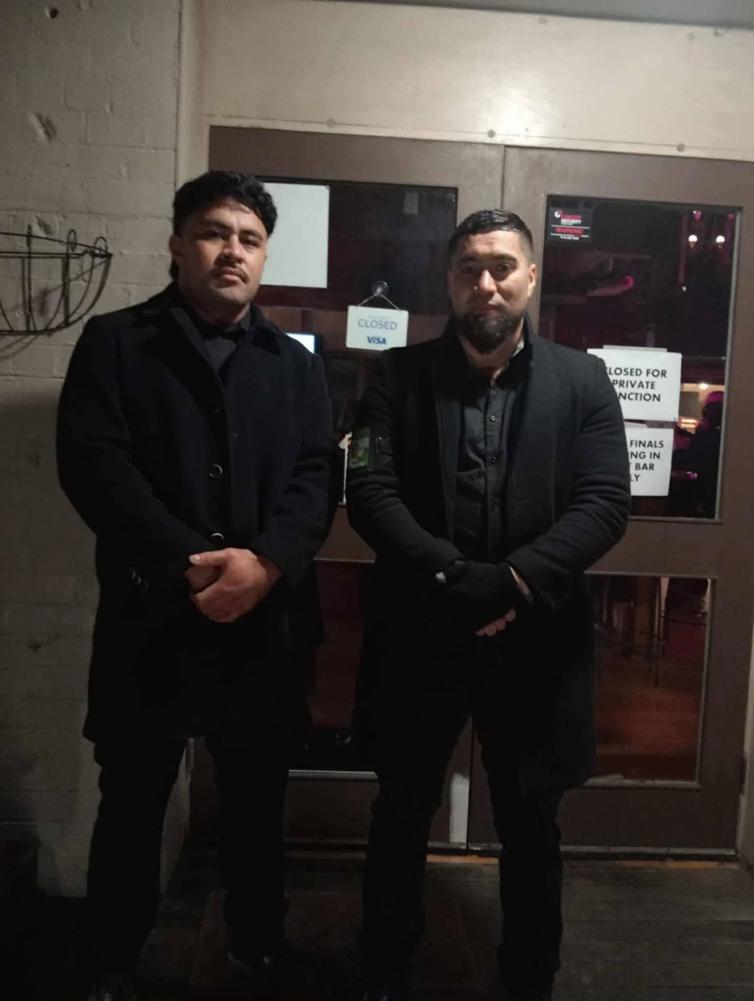 Two security officers standing outside a closed door at night, both wearing black jackets and gloves, with signs on the door indicating it is closed for private functions.